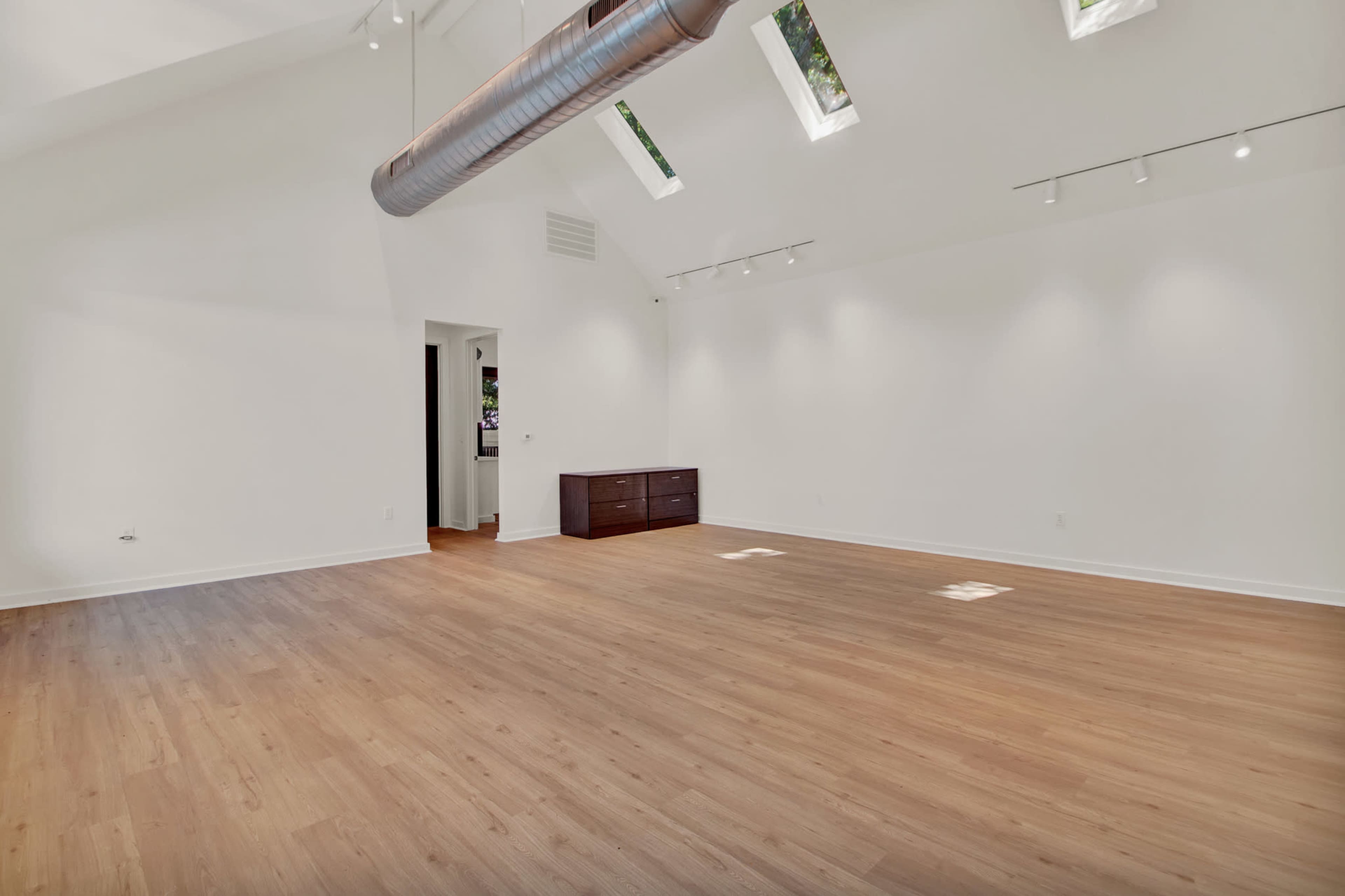 The image shows a spacious, empty room with high ceilings, wooden flooring, and a sleek light-colored wall.