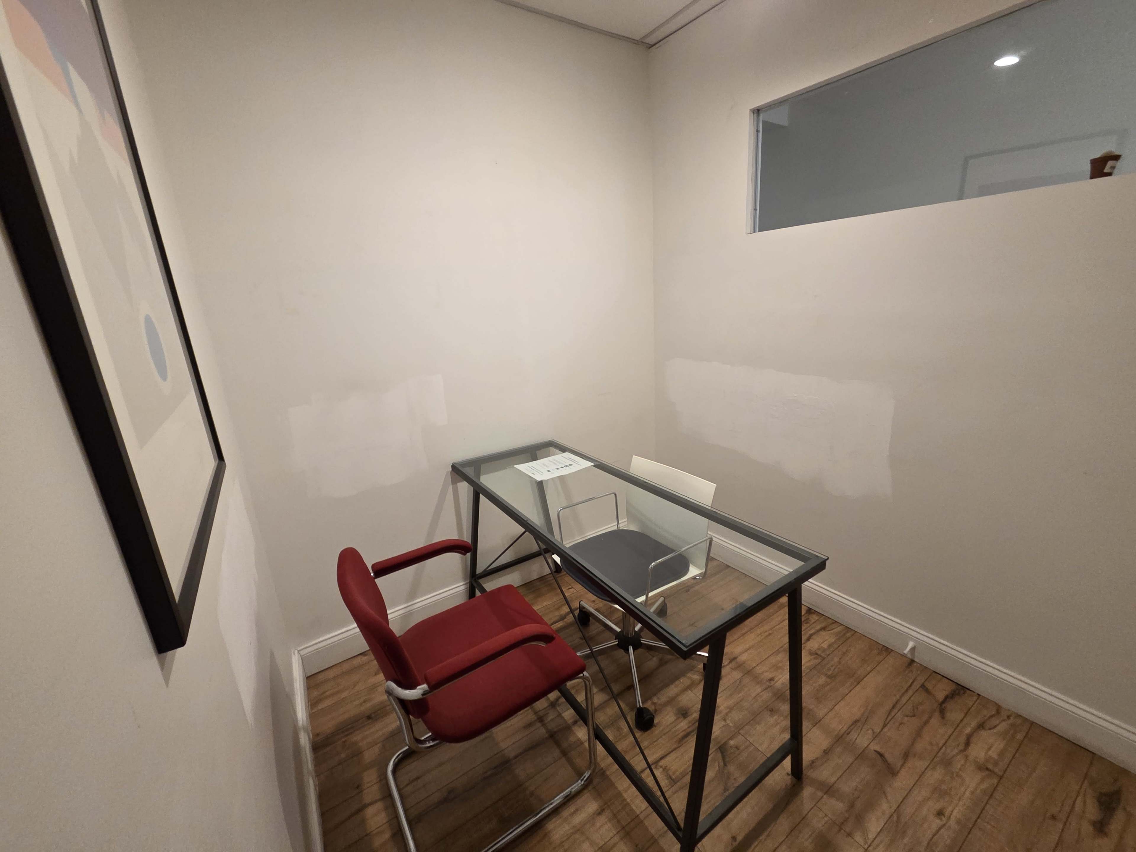 The image shows a small office space with a glass desk, a red chair, and a white wall with a small window.