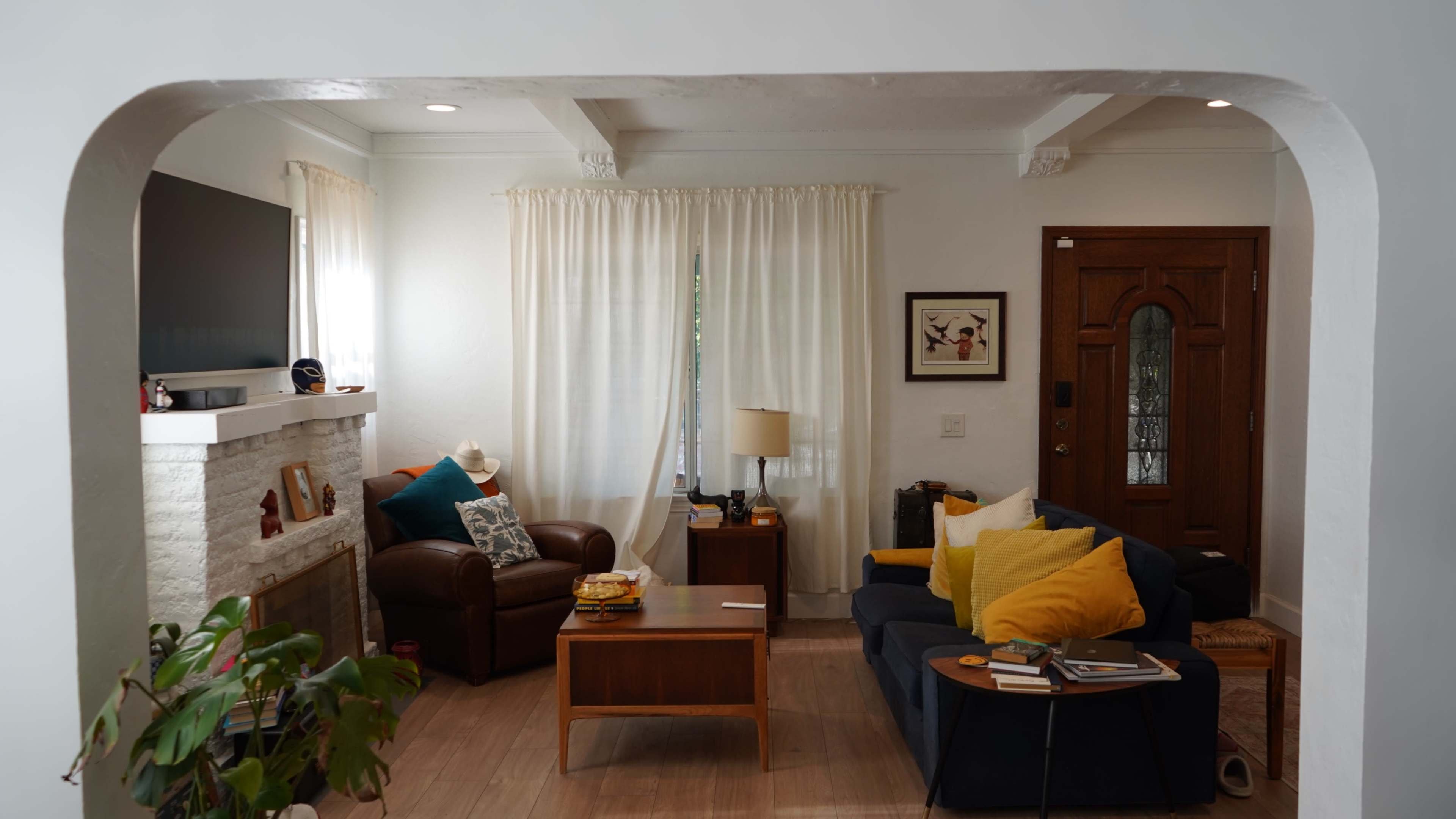 The image shows a cozy living room with a brown leather sofa, a blue sofa, a wooden coffee table, and a fireplace, complemented by light curtains and decorative cushions.