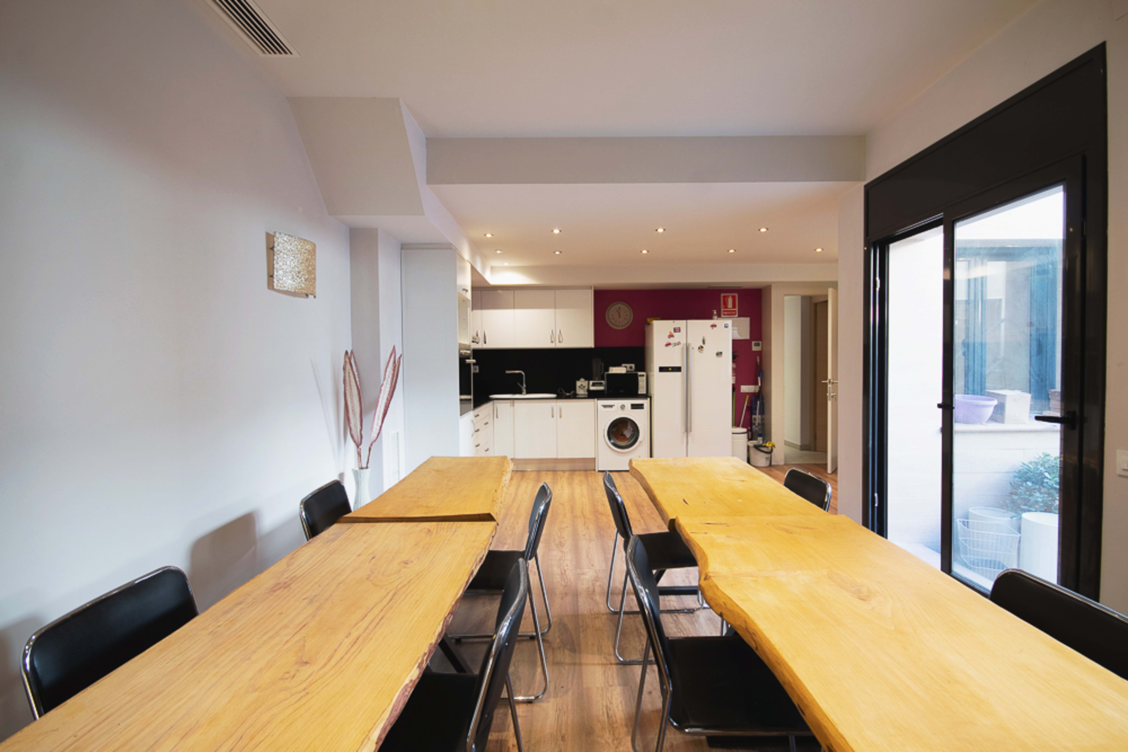 A spacious dining area features long wooden tables and black chairs, leading to a modern kitchen with white cabinetry and appliances.