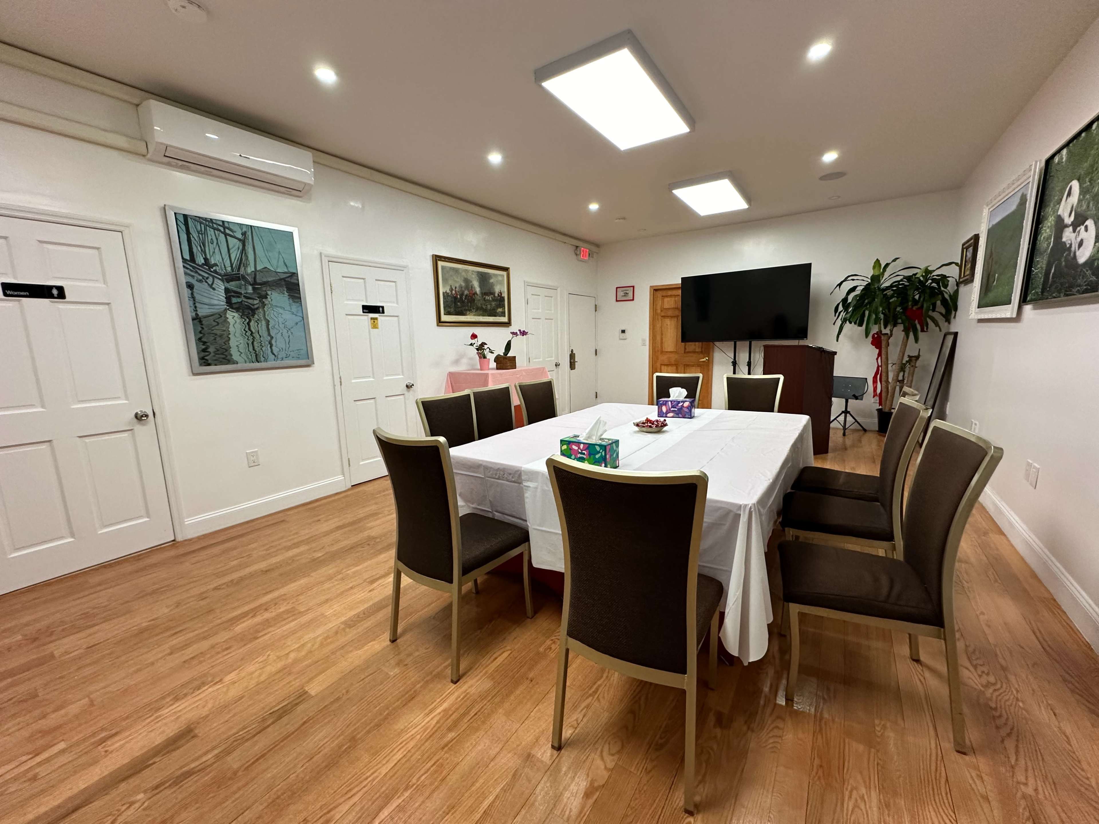The image shows a well-lit meeting room with a rectangular table surrounded by chairs, artwork on the walls, and a TV mounted in one corner.
