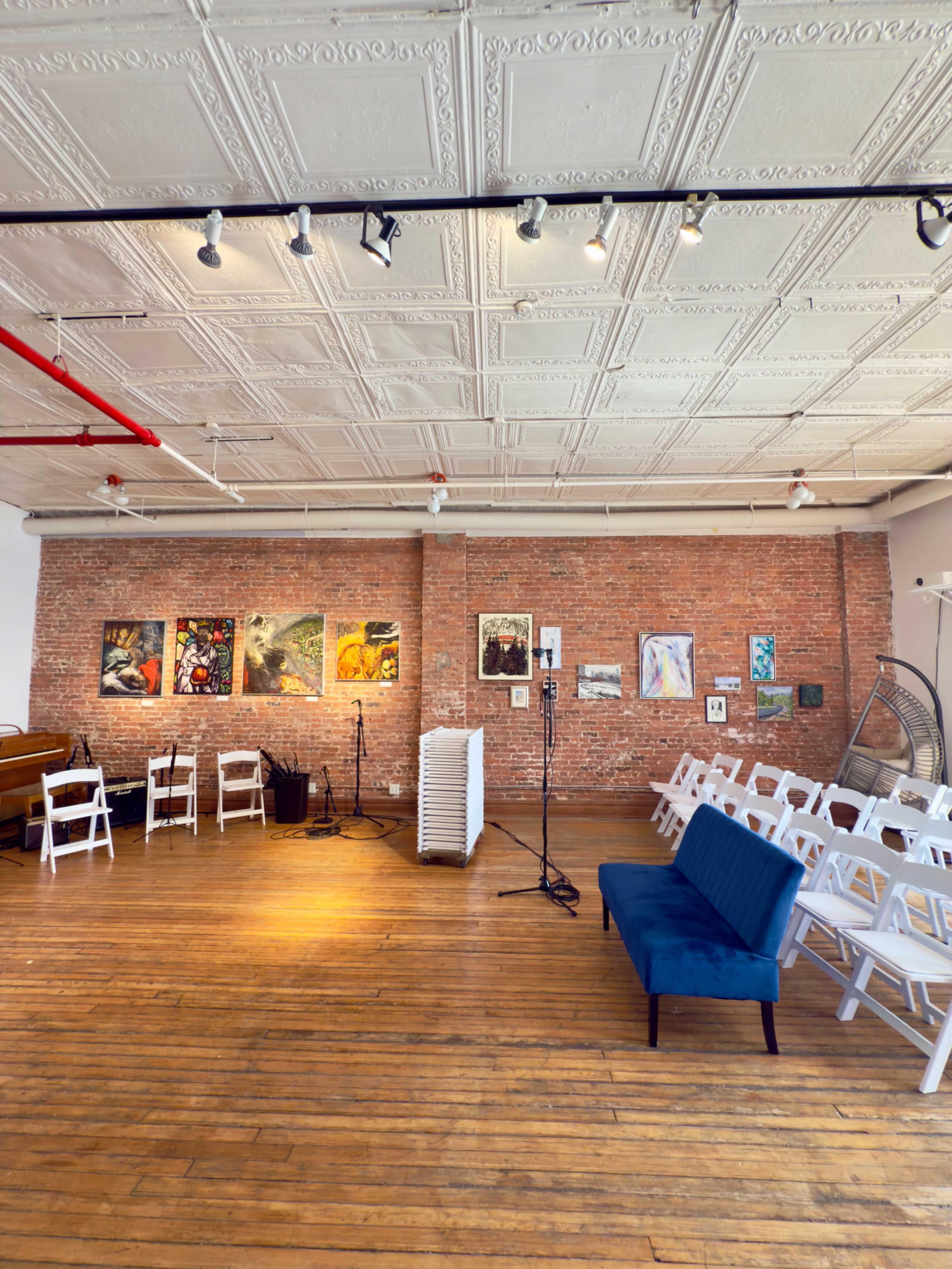 The image shows an art gallery with exposed brick walls, wooden flooring, and rows of white chairs facing a small stage with musical equipment and art pieces displayed on the walls.