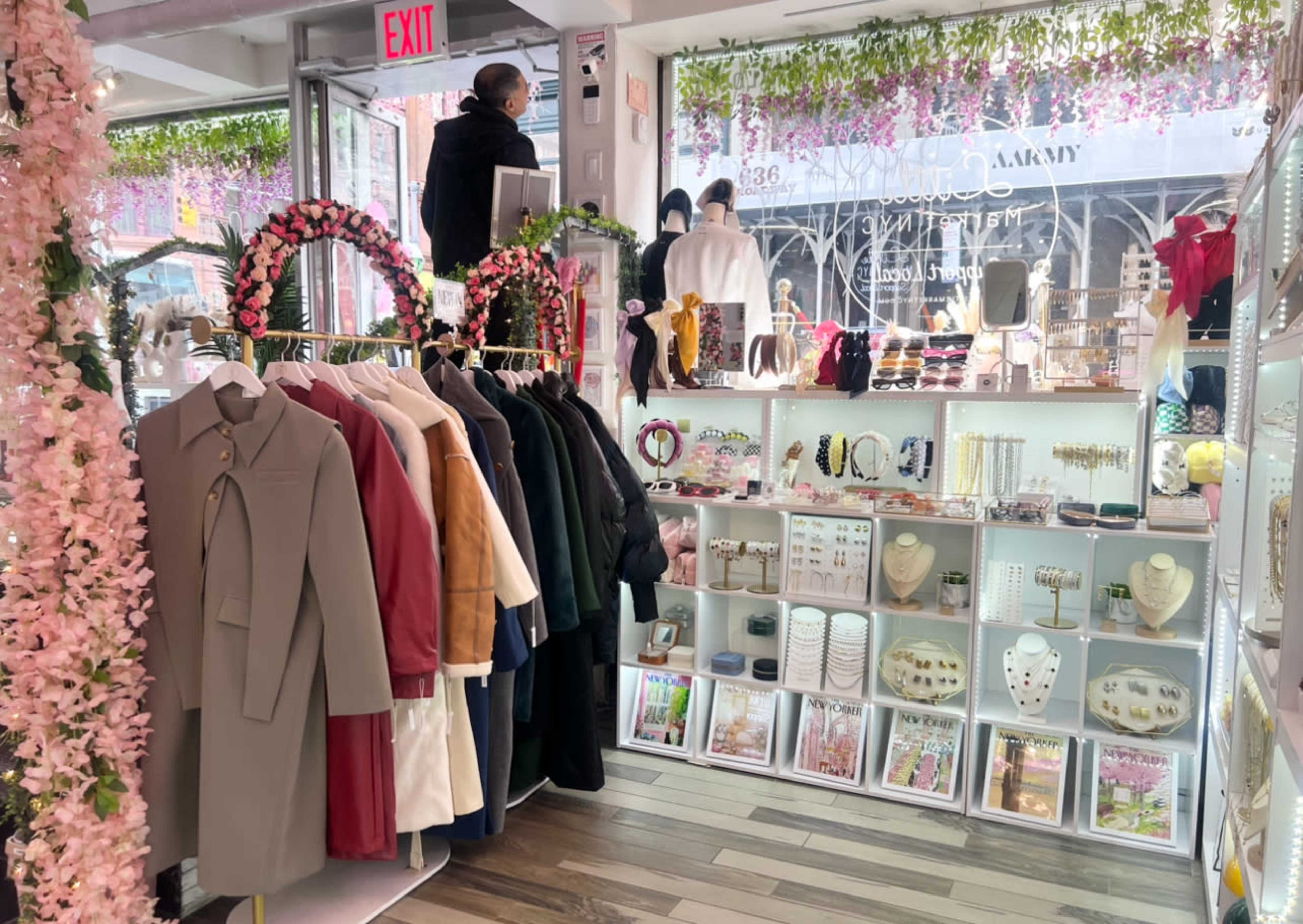 A boutique interior with clothing racks on the left and display shelves filled with accessories and jewelry on the right, alongside floral decorations in the shop windows.