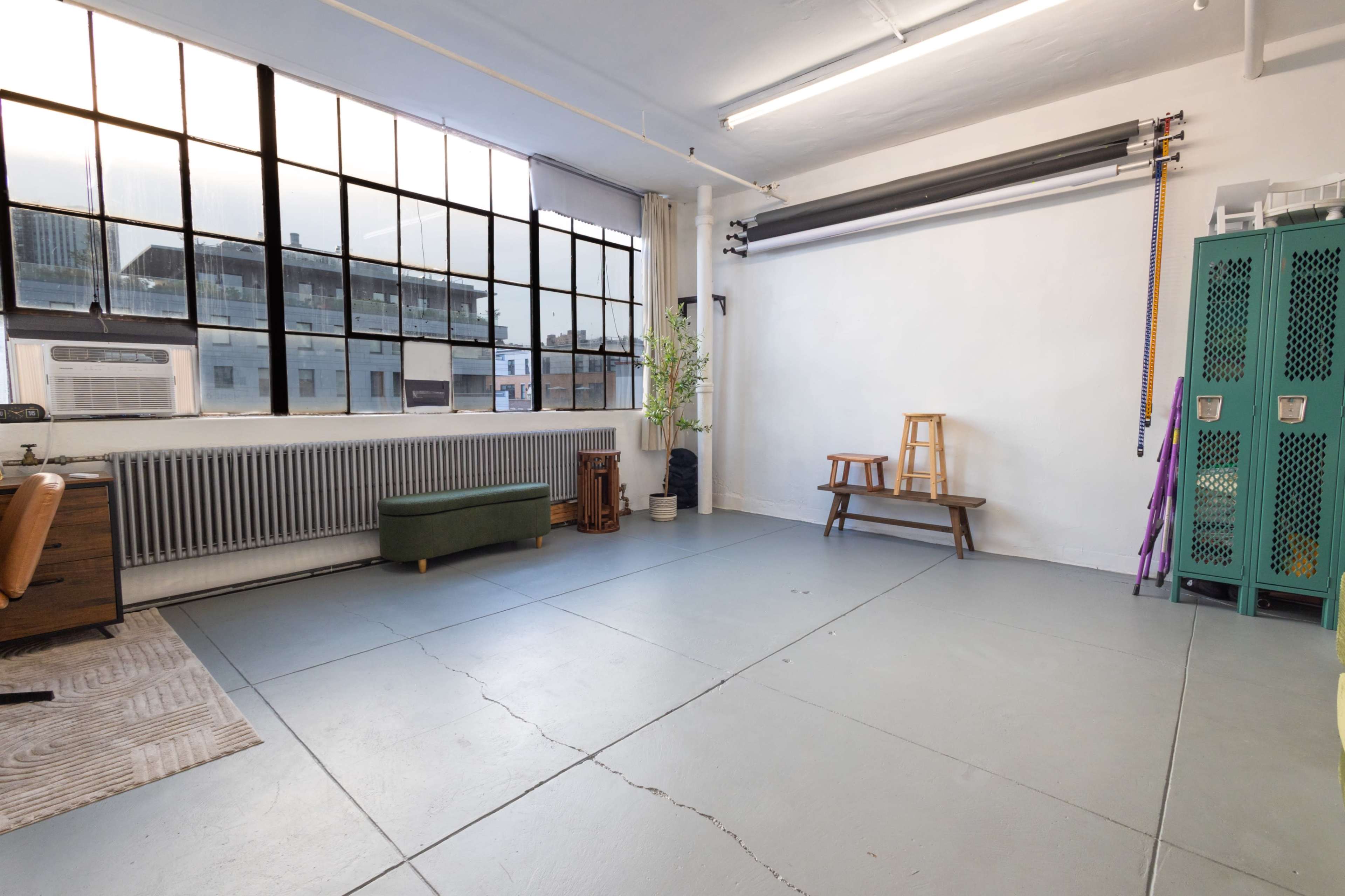 The image shows a spacious, well-lit studio with large windows, a green couch, a folding chair, and two green storage lockers against a white wall.