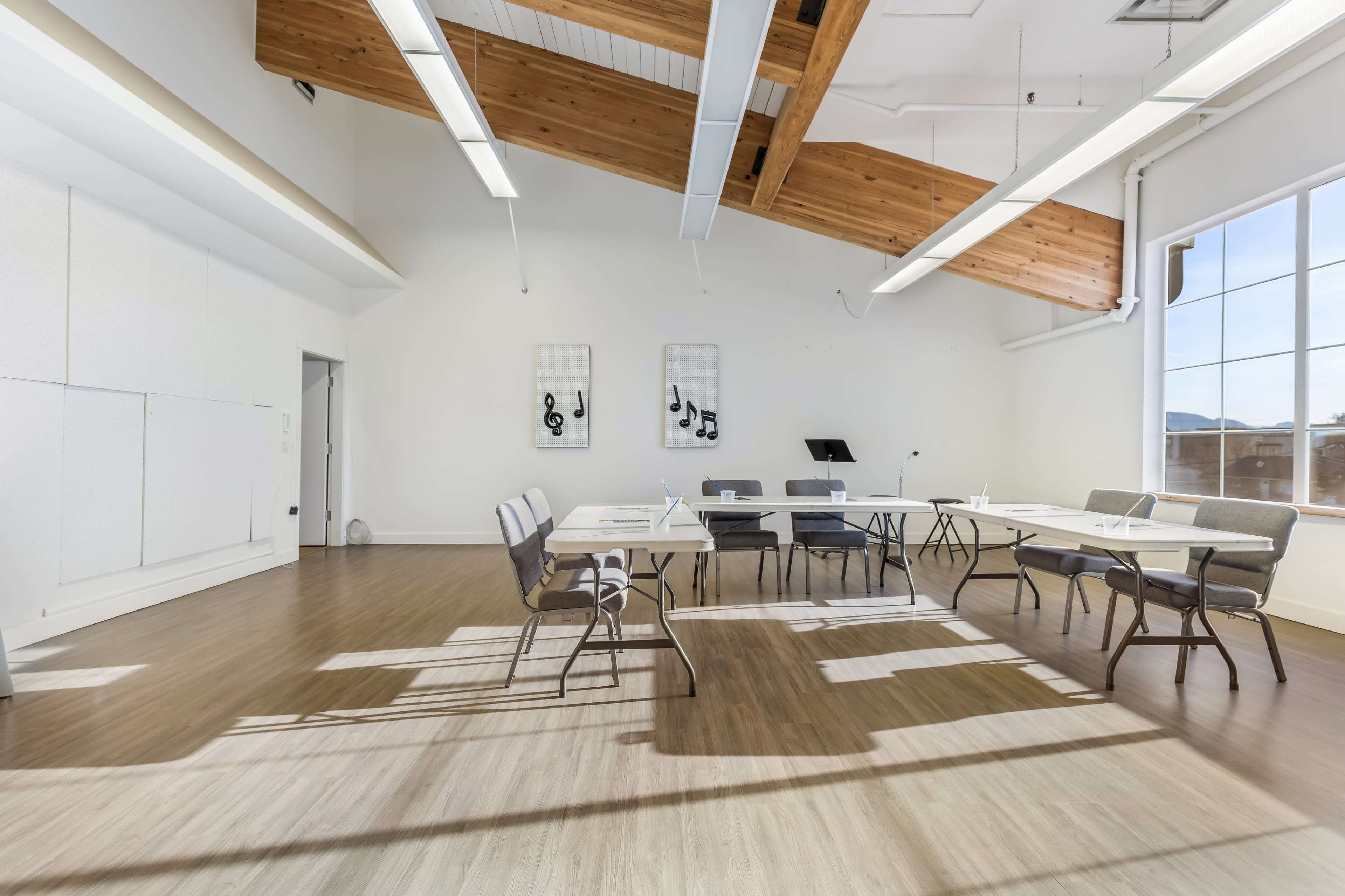 A spacious meeting room features large windows, wooden beams, and several tables arranged with chairs, illuminated by natural light.