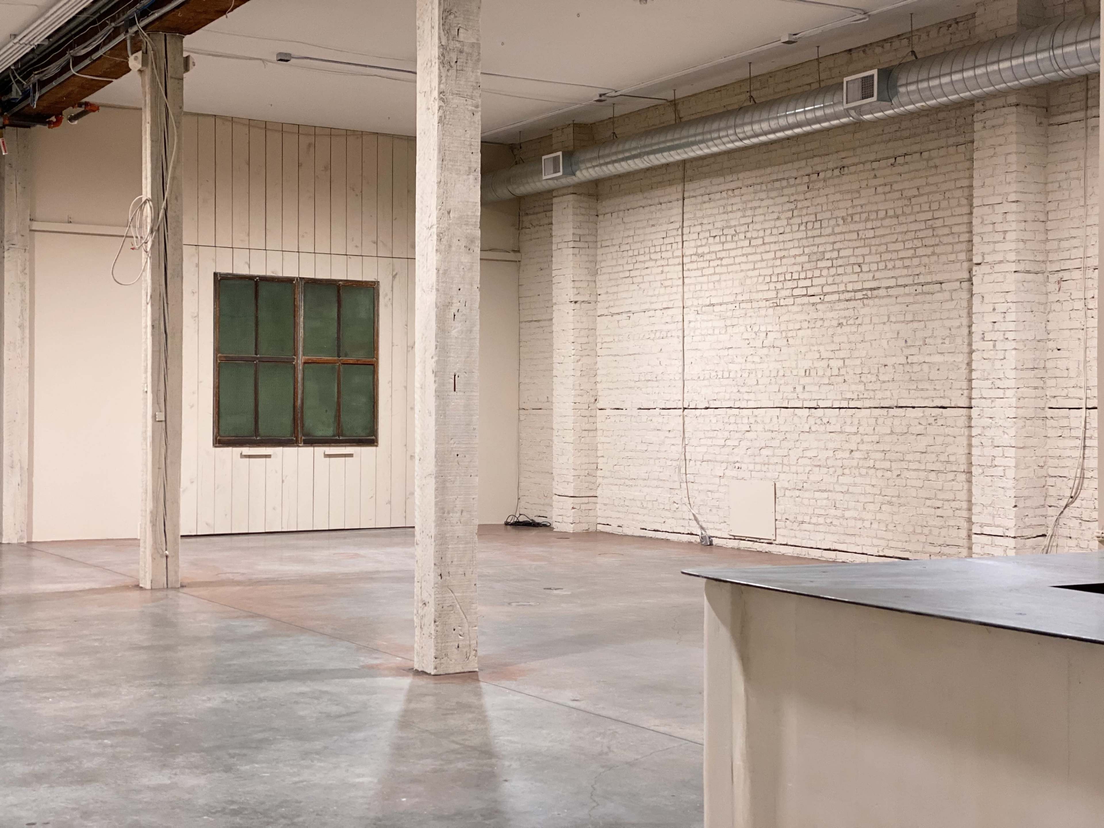 An empty industrial space with exposed concrete floors, white brick walls, wooden beams, and a window on one side.