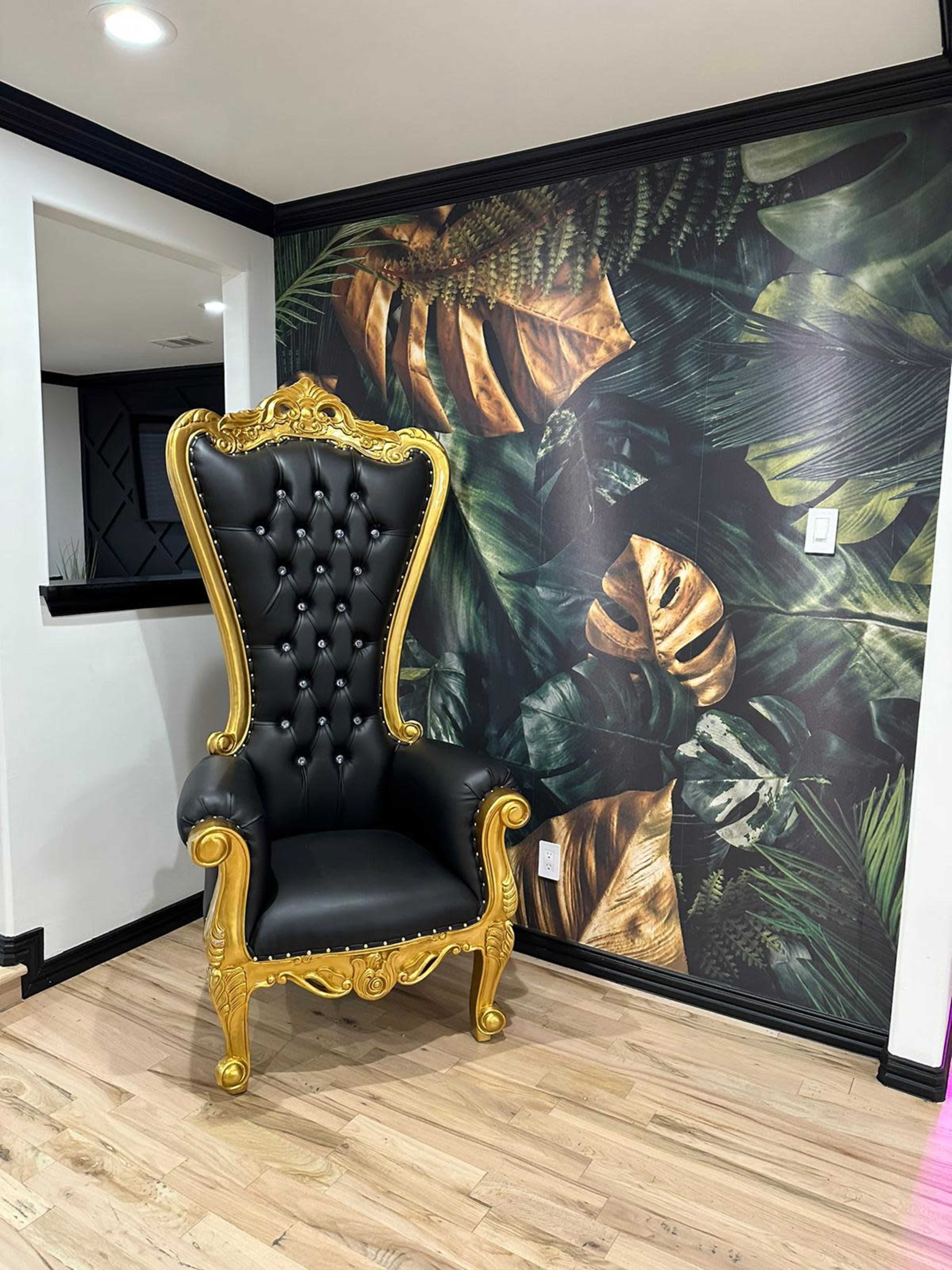 A tall, ornate black and gold throne chair is positioned against a wall adorned with tropical leaf wallpaper.