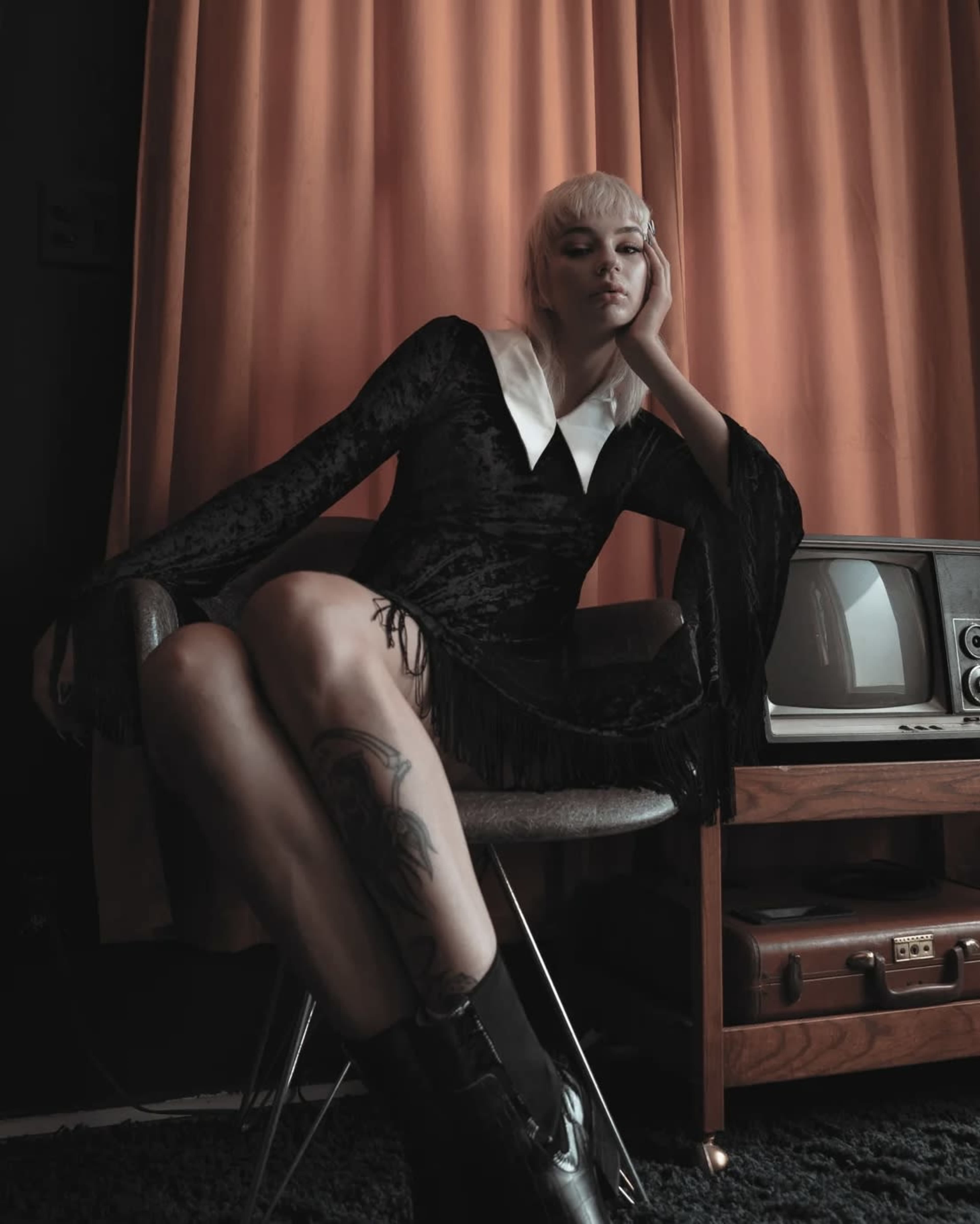 A person sitting in a chair, wearing a black dress with a white collar, in front of a vintage television and curtains.