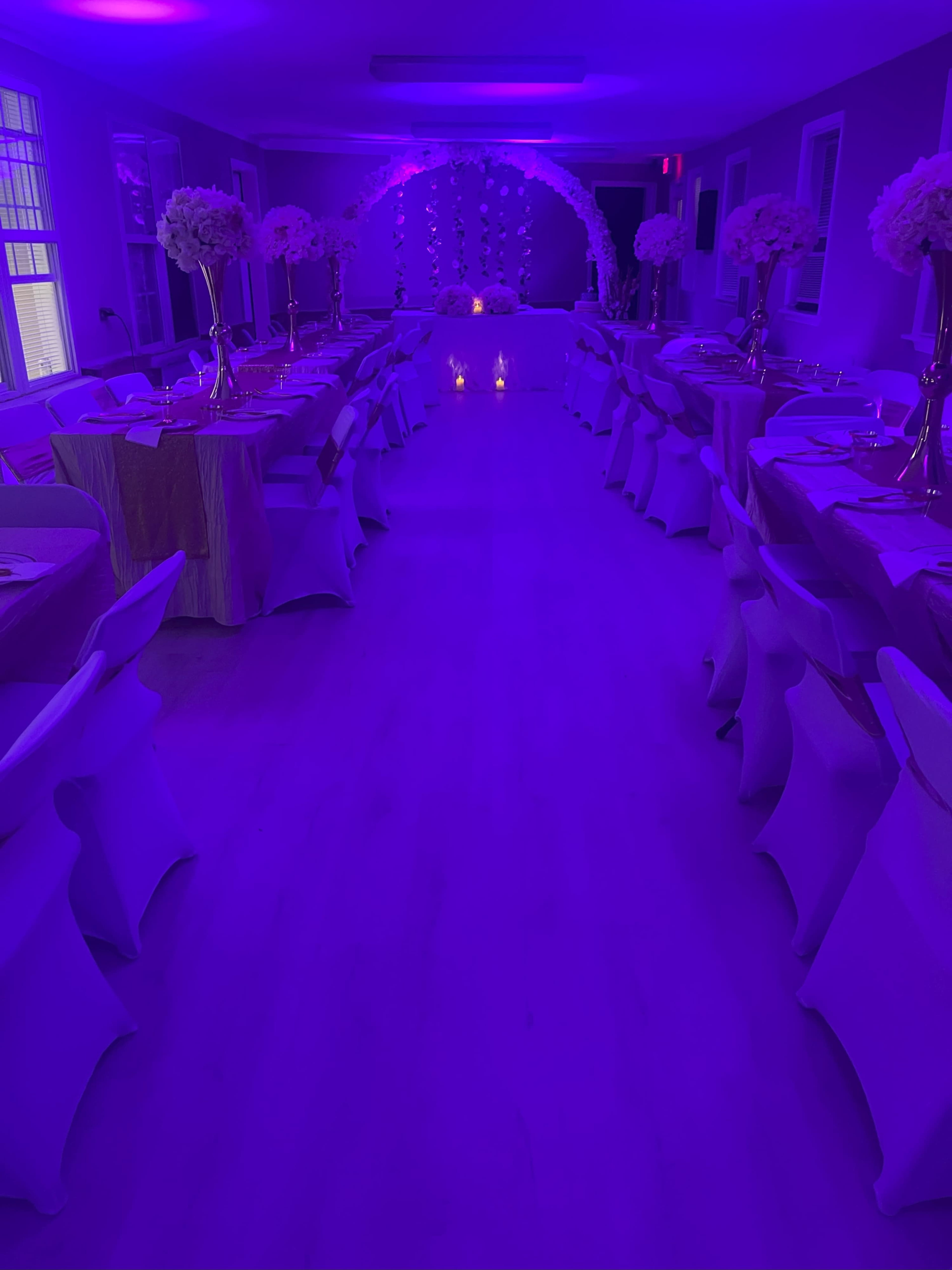 A banquet hall is decorated with purple lighting, featuring tables with elegant covers and floral centerpieces arranged along a central aisle.