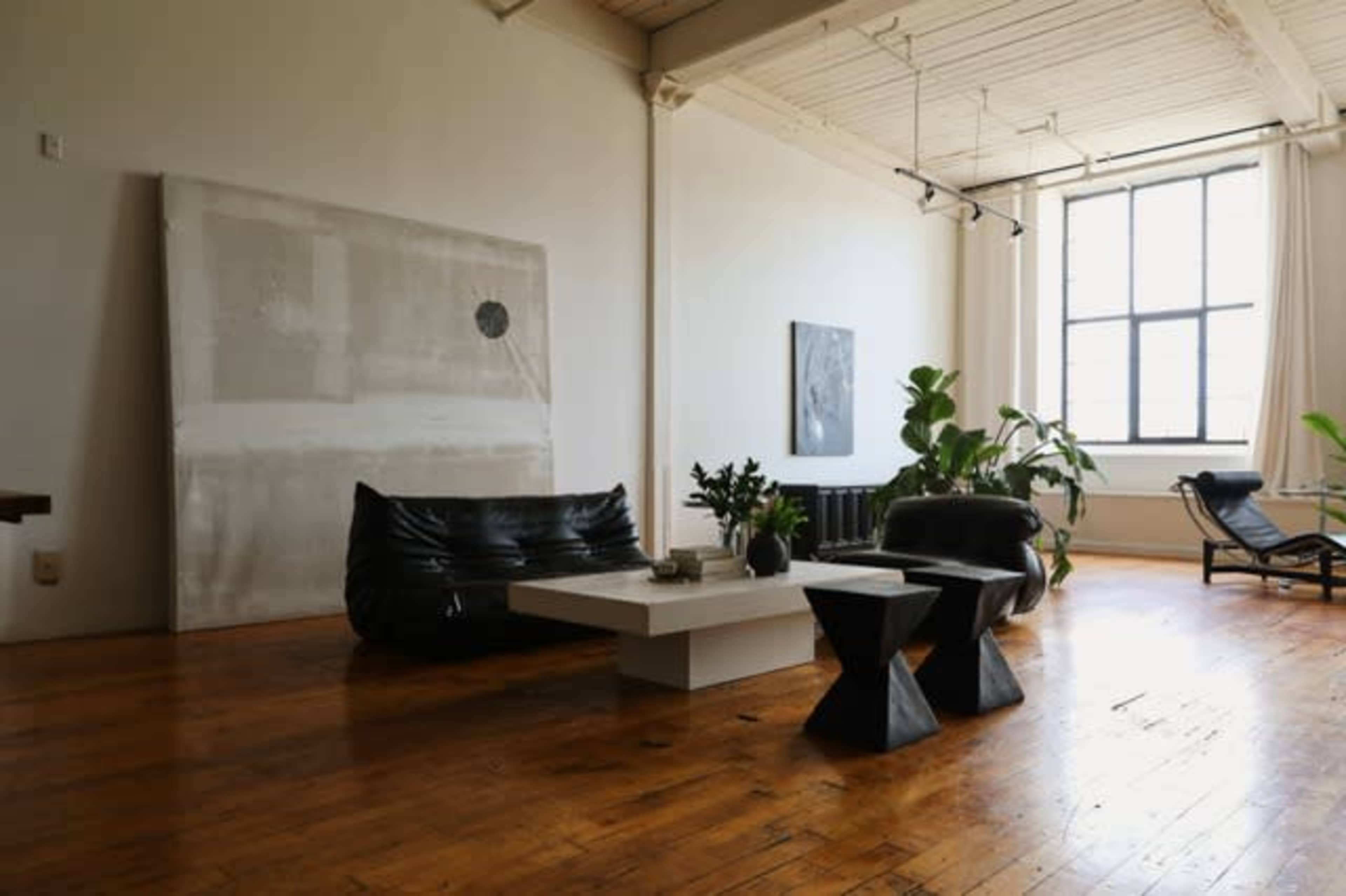 The image shows a modern, minimally furnished living space with a black bean bag chair, a white coffee table, and two dark sculptural stools, complemented by large windows and indoor plants.