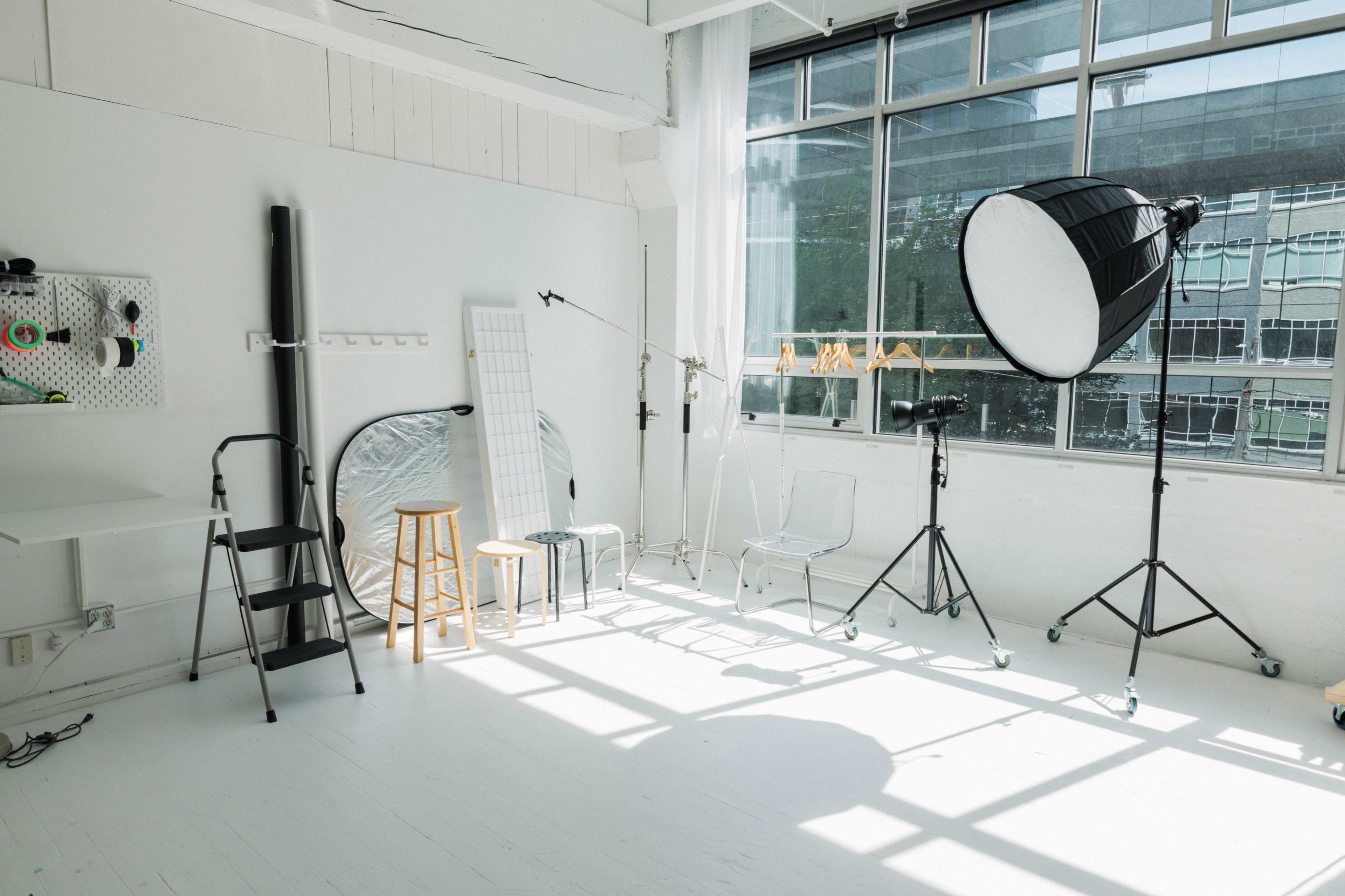 A well-lit photography studio featuring various equipment, including a large softbox, a reflector, and several chairs against a backdrop of large windows.