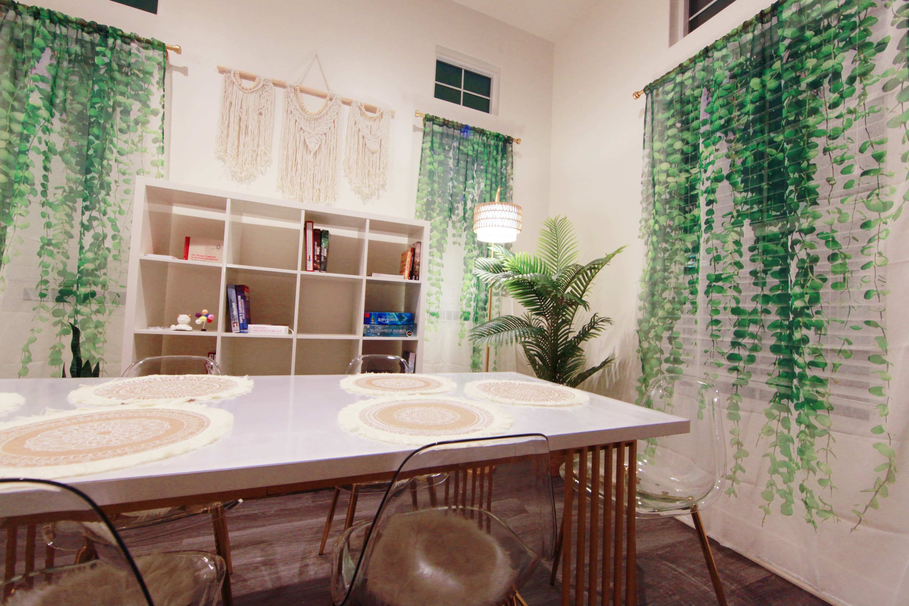 The image shows a well-lit room featuring a white table with placemats, clear chairs, a bookshelf, and curtains adorned with leafy patterns.