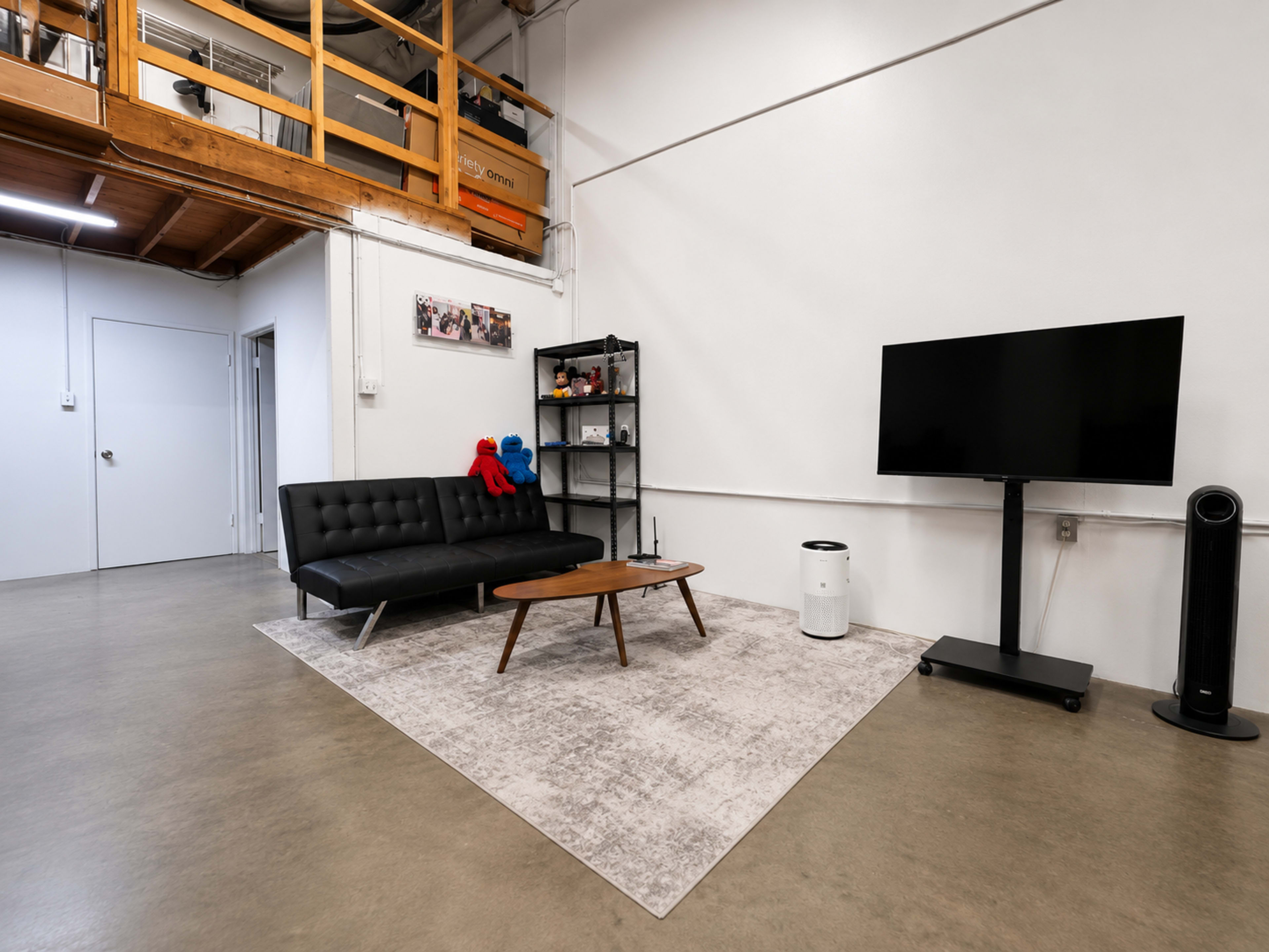 The image shows a modern living room featuring a black sofa, a wooden coffee table, a shelf with decorative items, and a flat-screen television mounted on a stand, all situated on a light-colored rug in a spacious area with exposed wooden beams above.
