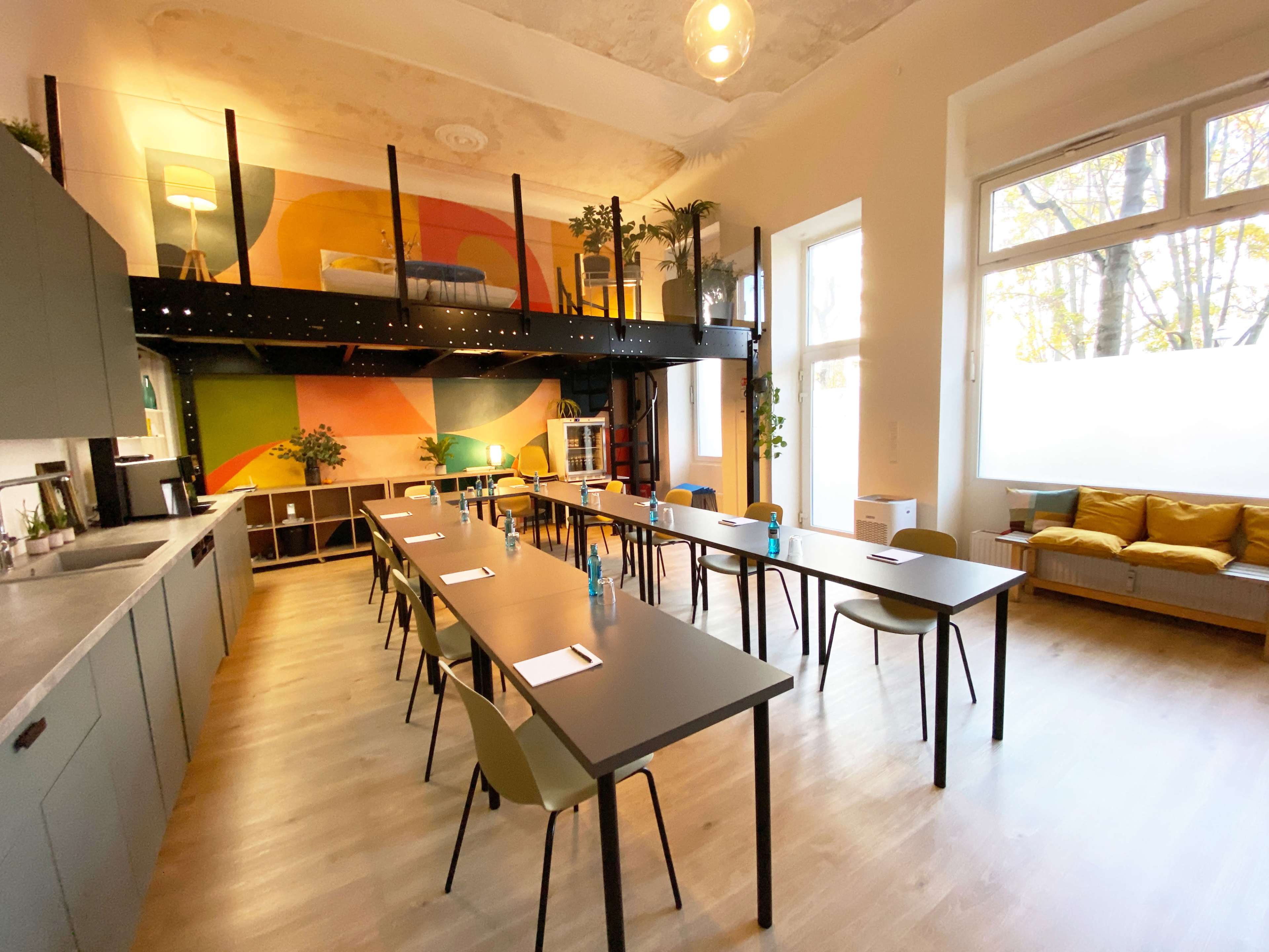 A spacious room features long tables arranged in rows, with a kitchenette and colorful wall art, illuminated by natural light from large windows.