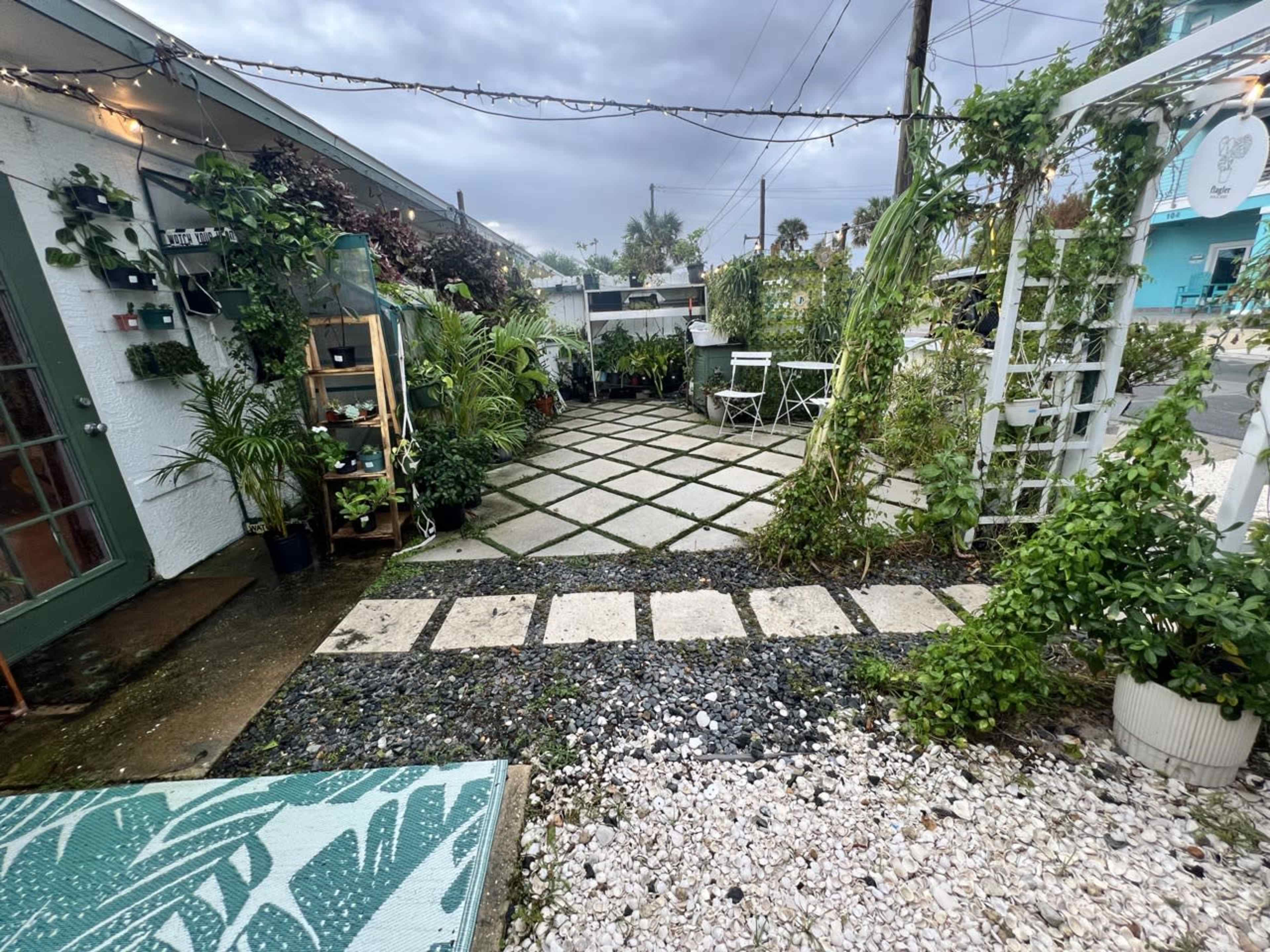 The image shows a landscaped outdoor area featuring a checkerboard pattern of pavers and gravel, bordered by plants and seating arrangements.
