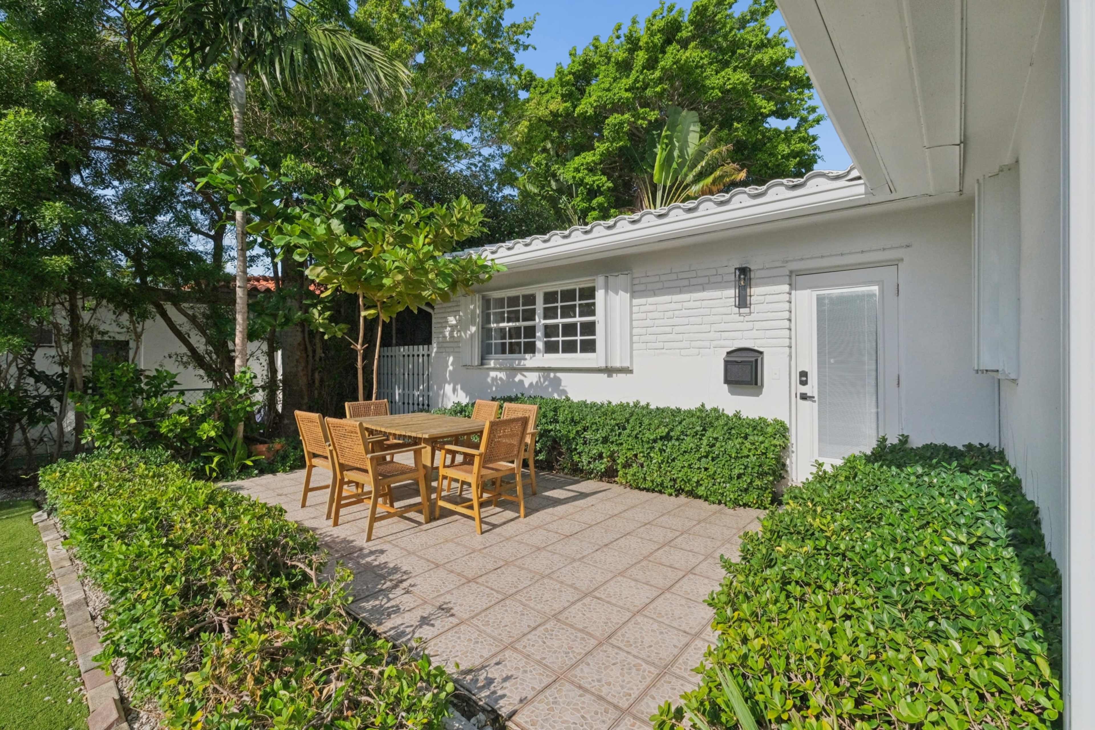 The image shows a brick-paved patio surrounded by greenery, featuring a wooden dining table with four chairs.