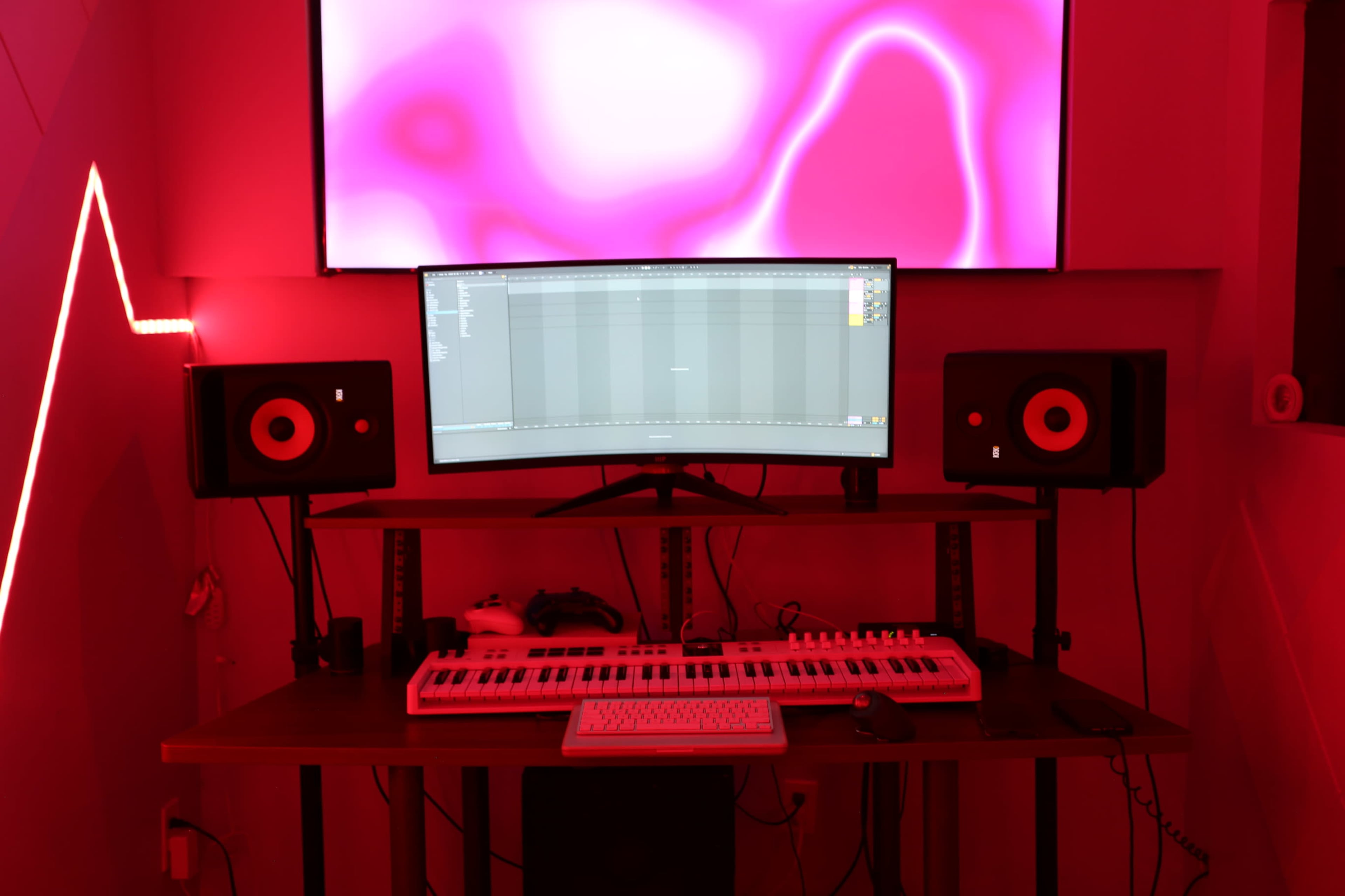 The image shows a music production setup featuring a desk with a computer monitor, studio monitors on either side, a MIDI keyboard, and a colorful backdrop illuminated in red.