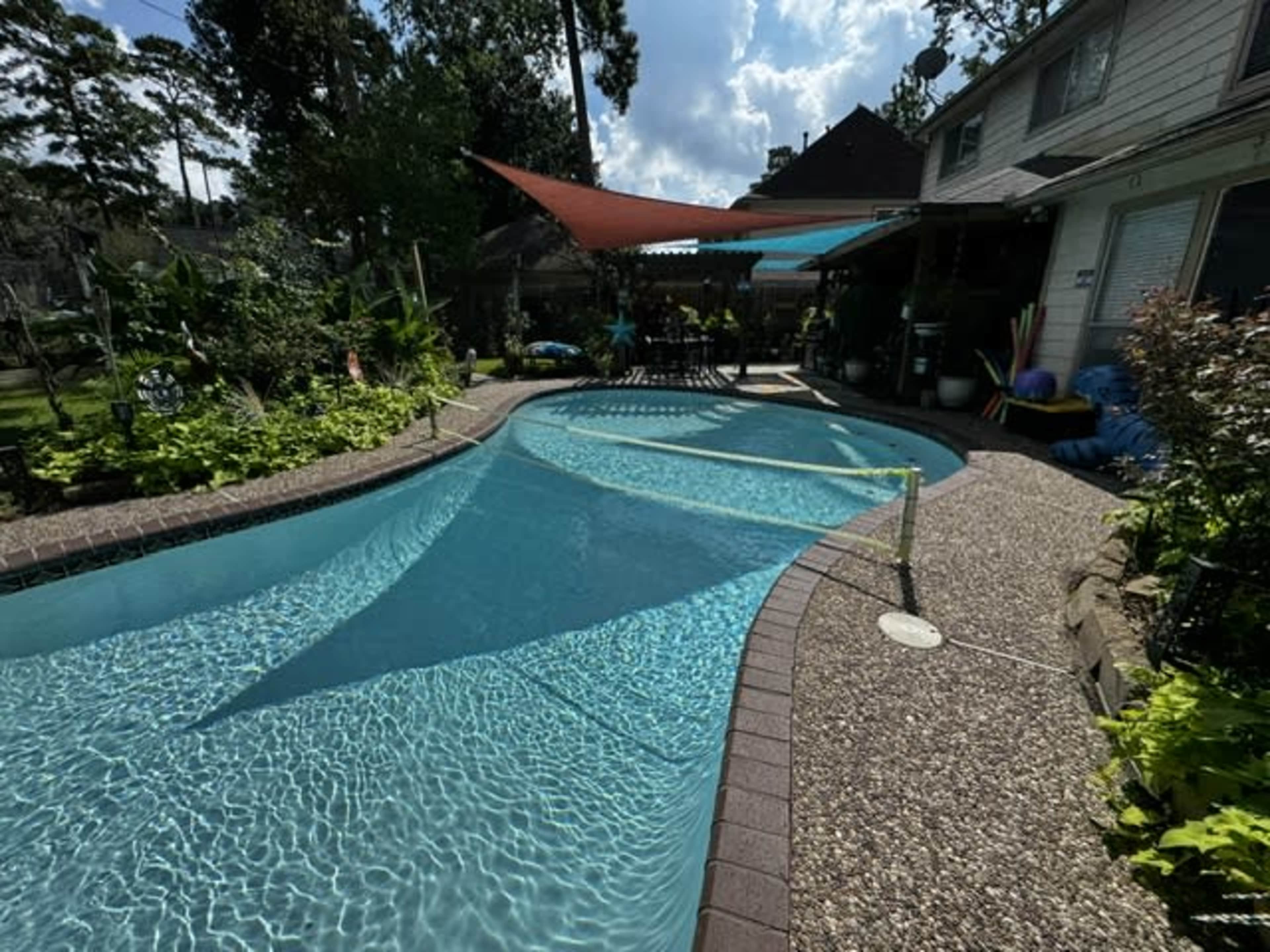 The image shows a residential pool surrounded by lush greenery and a shaded seating area with colorful pool accessories nearby.