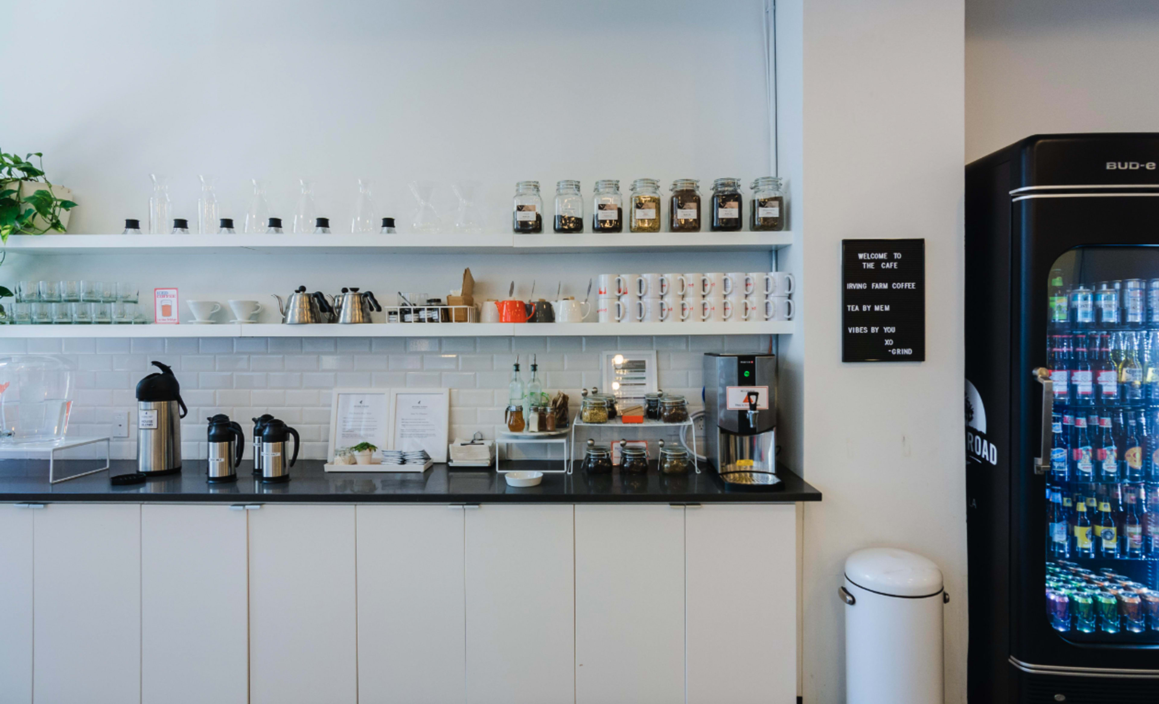 A coffee bar with a neatly arranged countertop featuring jars of ingredients, mugs, brewing equipment, and a vending machine against a white wall.