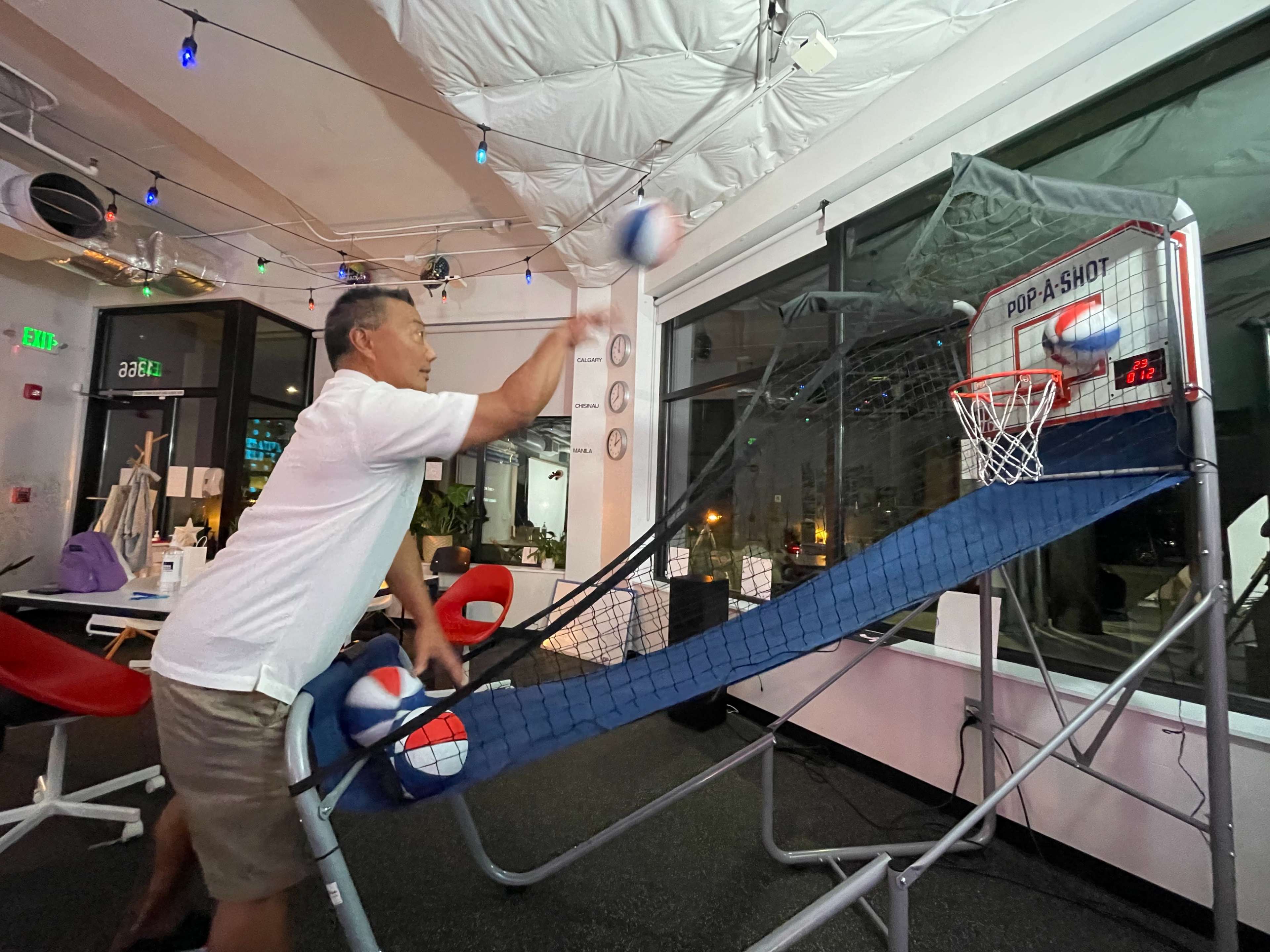 A man is playing basketball at an arcade game setup, shooting balls into a netted hoop in a brightly lit indoor space.