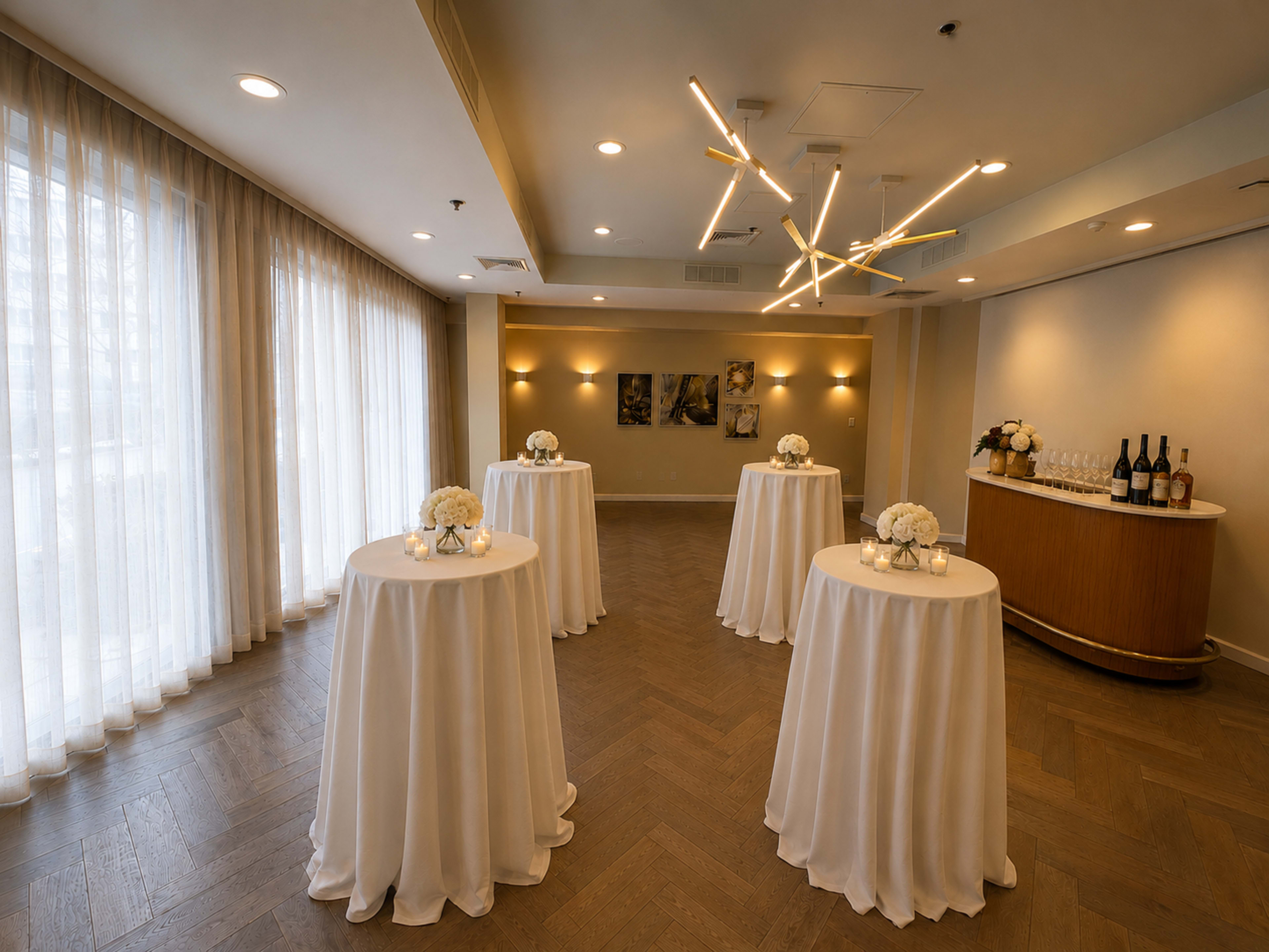 The image shows a well-lit event space featuring five round tables draped in white cloth, each adorned with a floral centerpiece, and a bar area in the background with bottles.