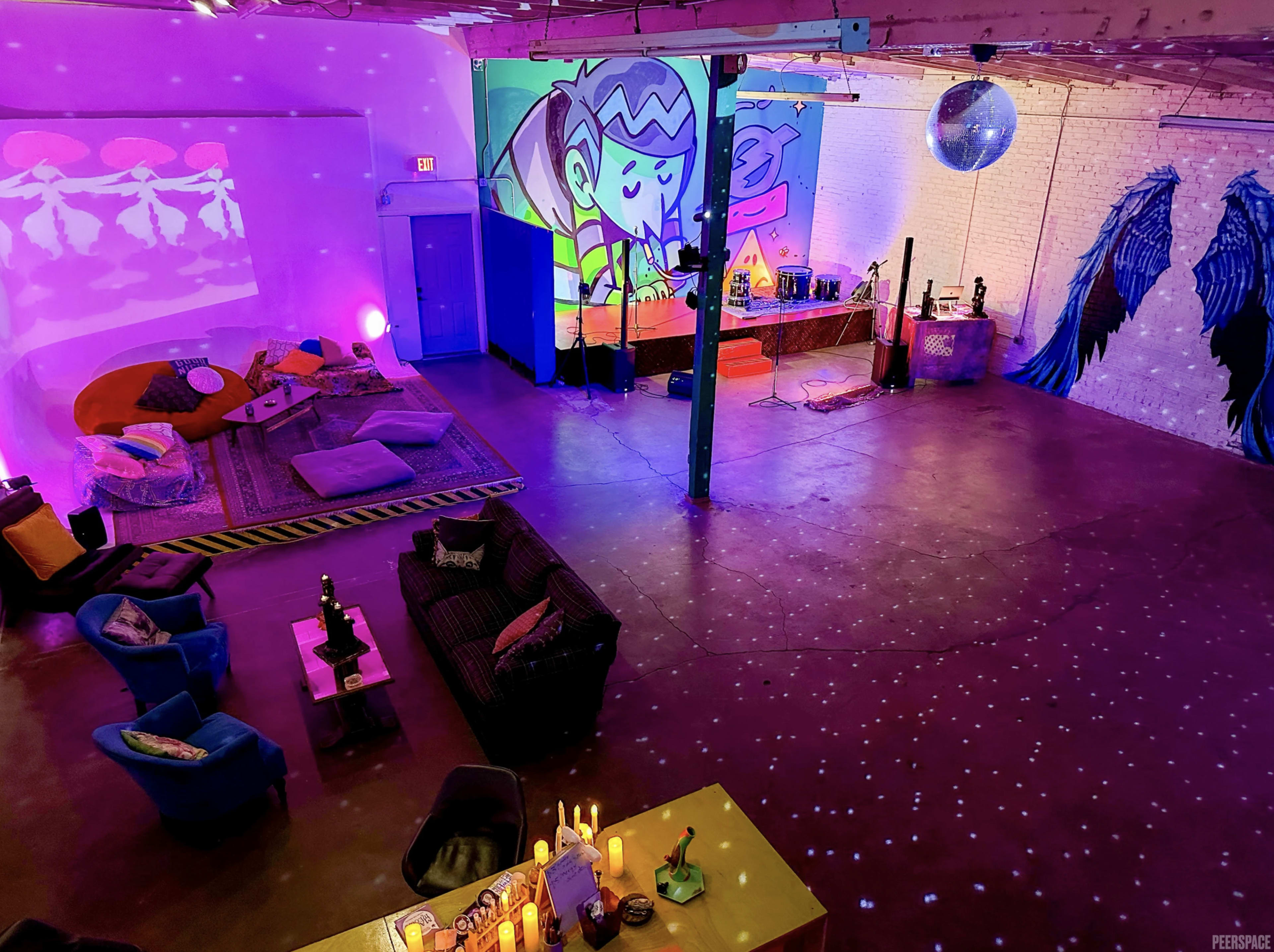 The image shows a spacious, brightly colored room with eclectic furniture, vibrant murals on the walls, and a disco ball hanging from the ceiling.