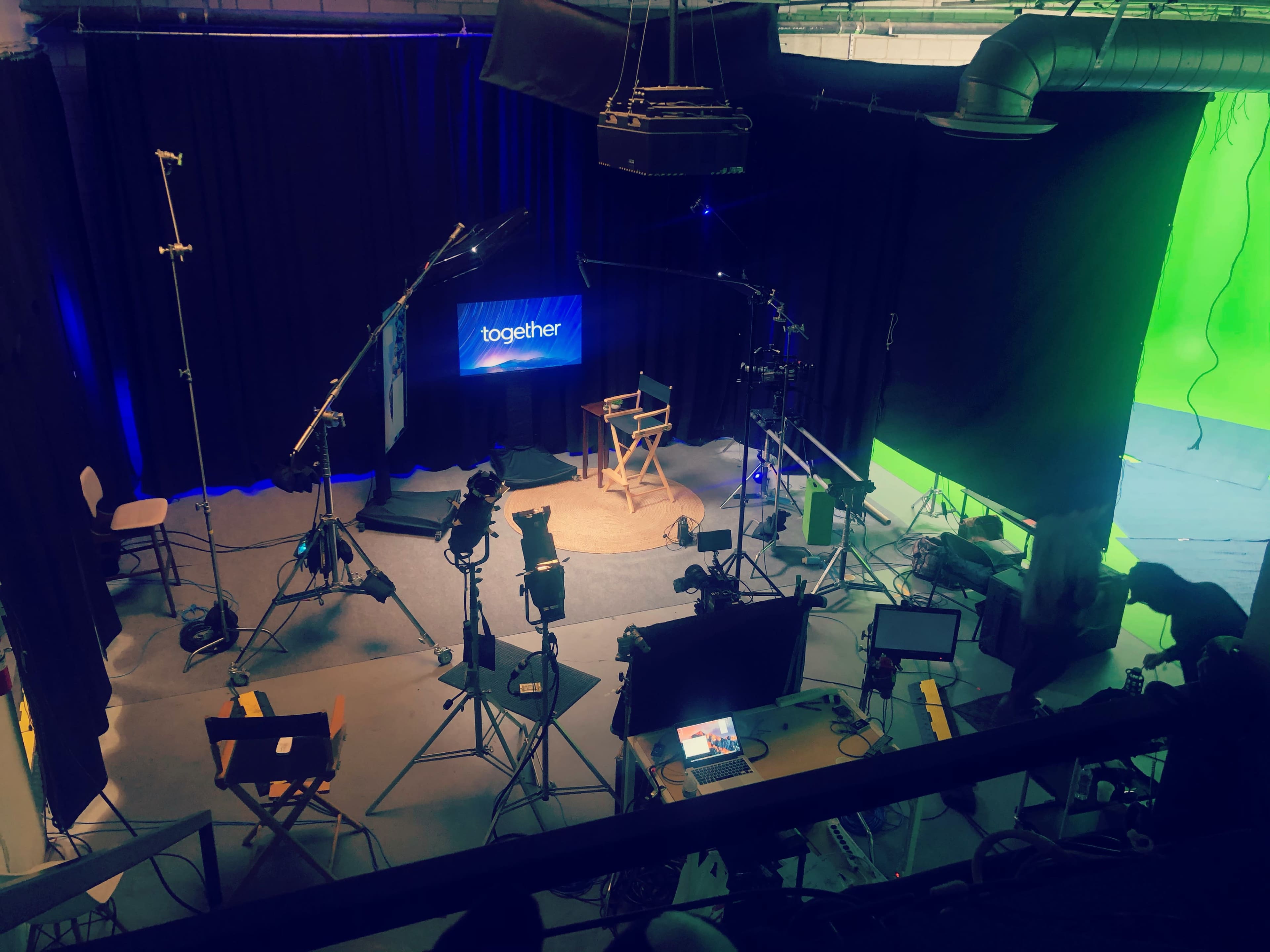 The image shows a film studio setup featuring multiple cameras, lighting equipment, and a green screen area, with a director's chair and screens positioned in the center.