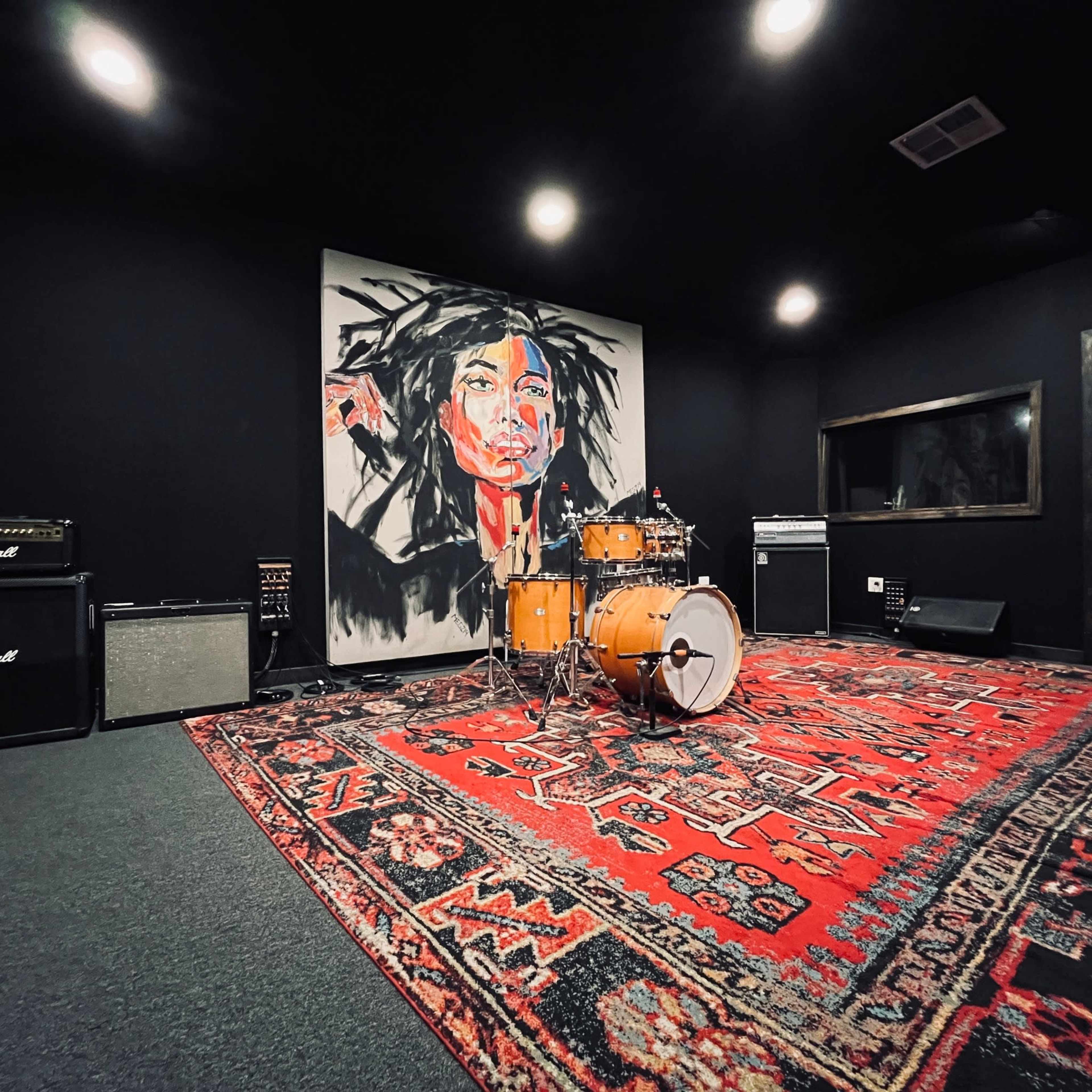 The image shows a rehearsal studio featuring a drum set in front of a large mural of a person's face, surrounded by amplifiers and a patterned rug on the floor.