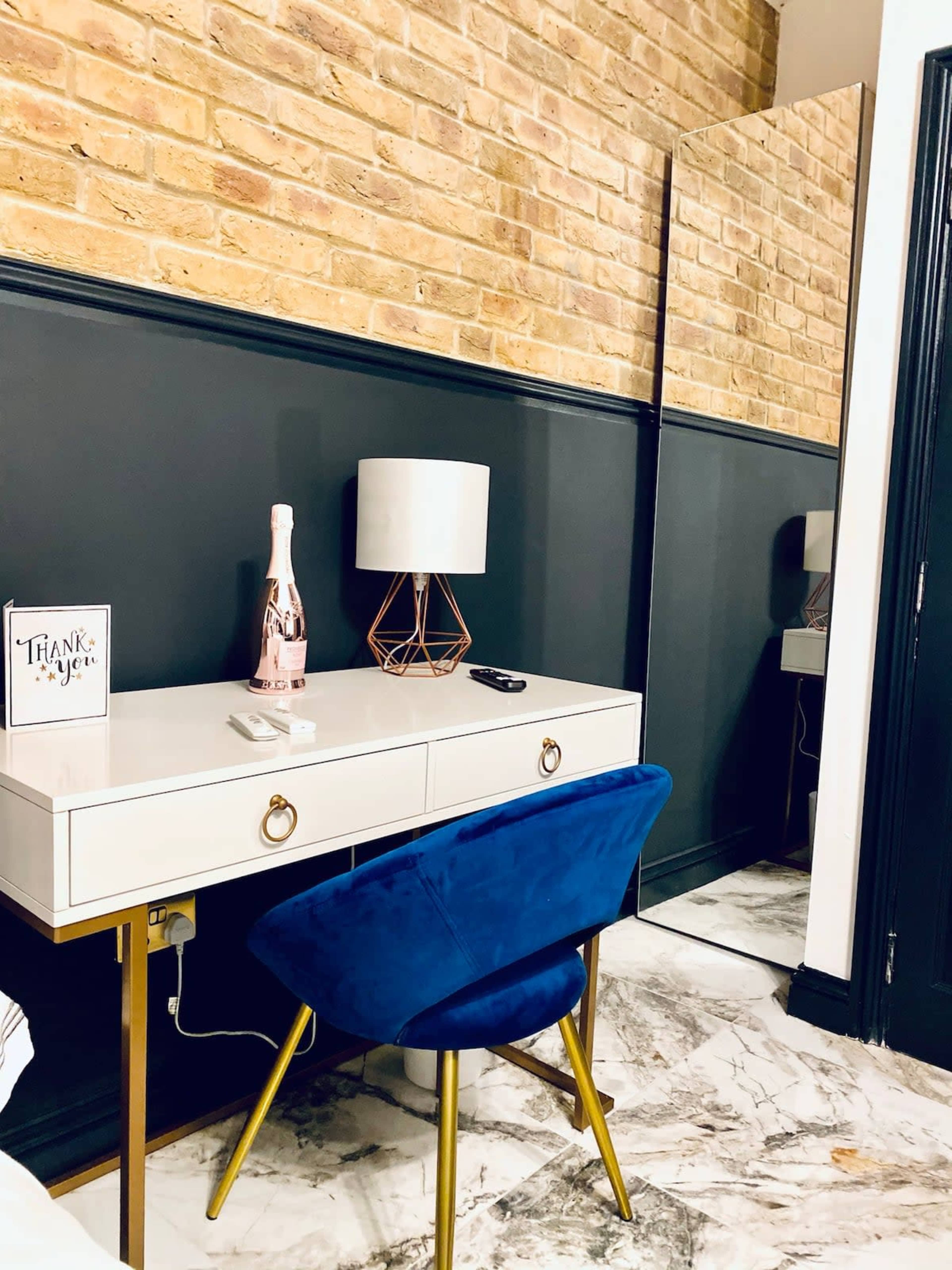 A desk with a blue chair, a lamp, and a decorative item is positioned against a wall with exposed brick and a large mirror on one side.