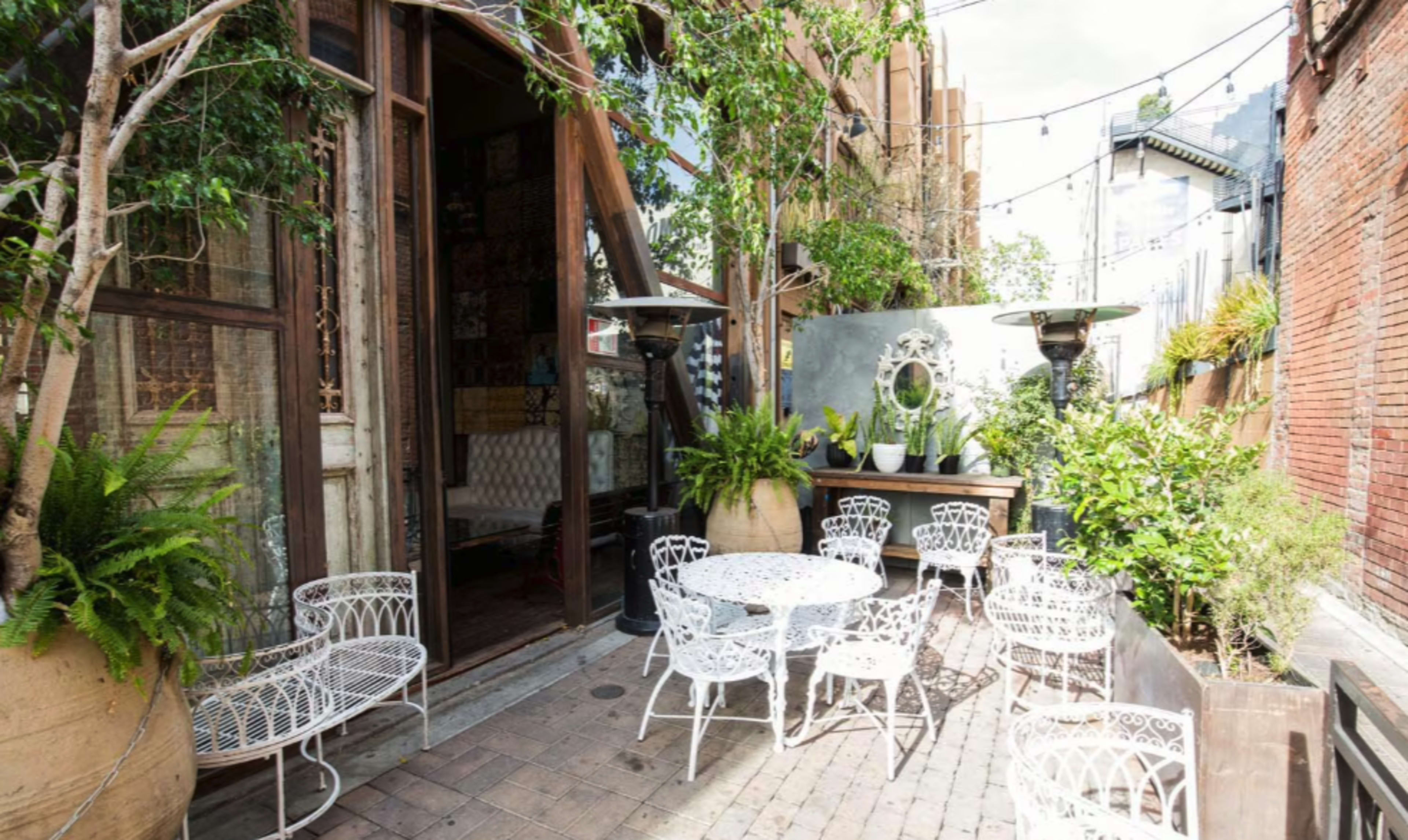 The image shows a small outdoor seating area with white wrought iron furniture, potted plants, and a wooden structure in the background.