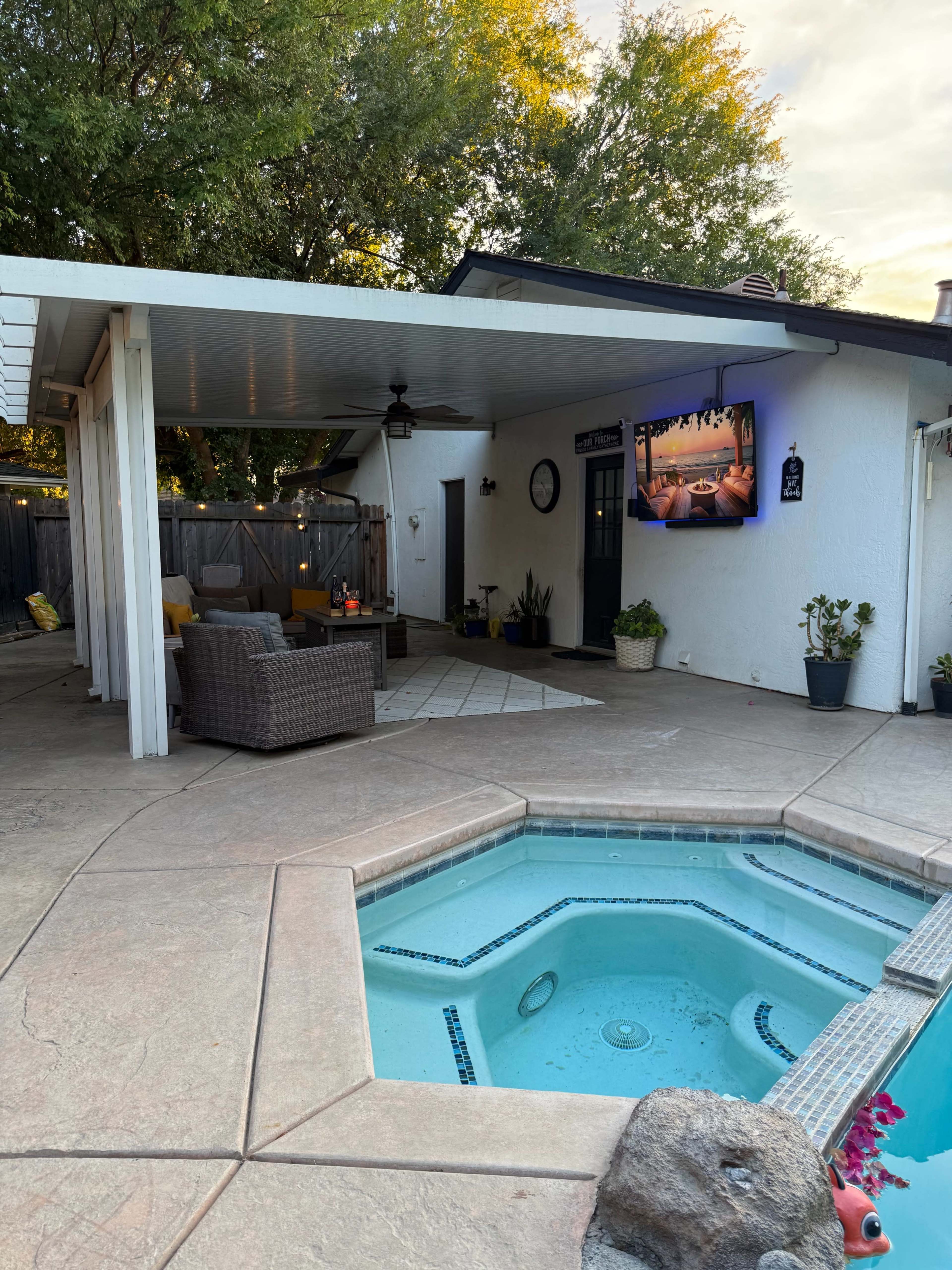 The image shows a backyard patio area with a hot tub, outdoor seating, and a mounted television on the wall, surrounded by greenery.