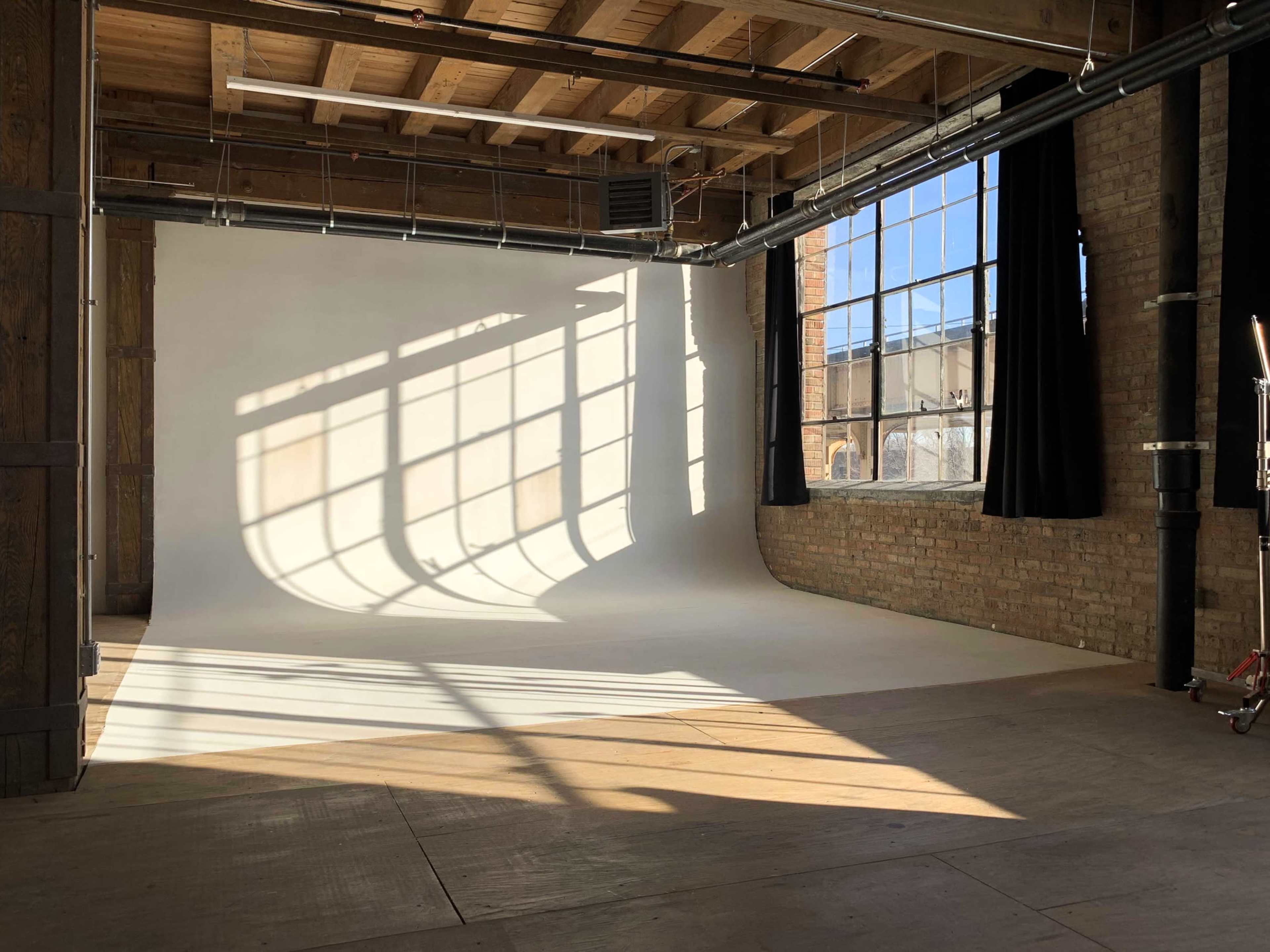 The image shows an empty photography studio with a white backdrop and sunlight casting shadows on the floor and wall from a large window.