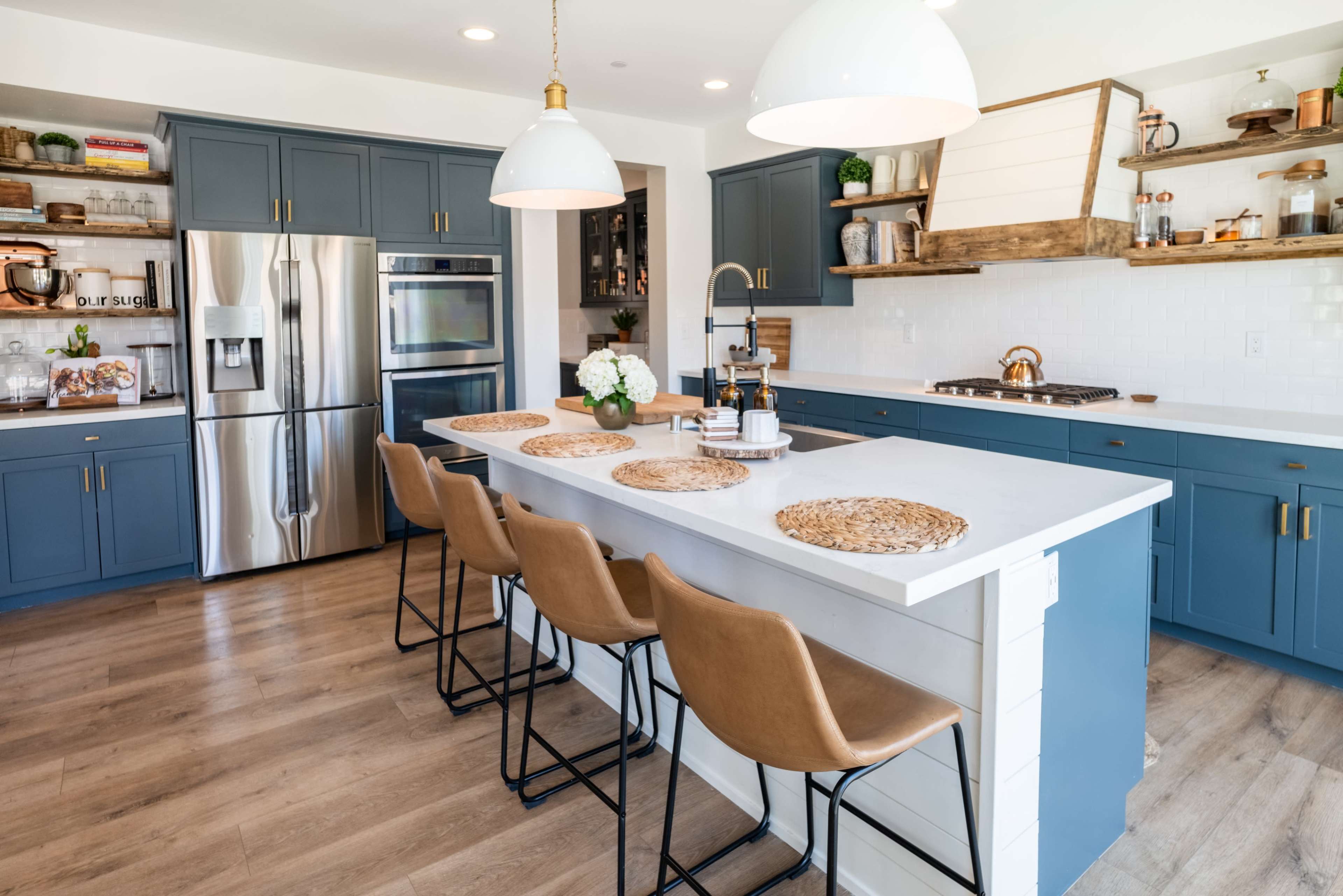 A modern kitchen features blue cabinets, stainless steel appliances, a white countertop, and four brown bar stools along a spacious island.
