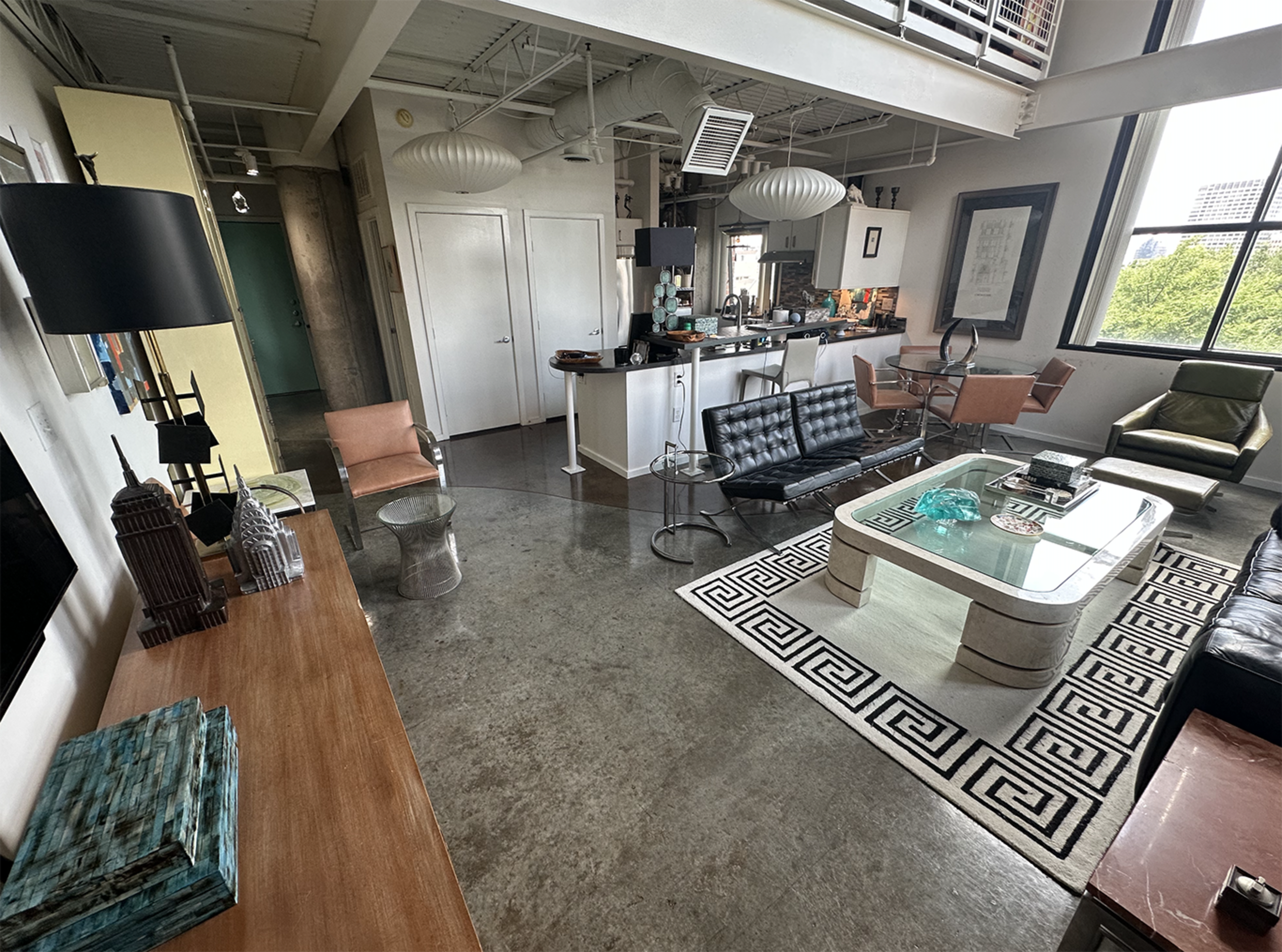 The image shows a modern, open-concept living area with a mix of furniture including a glass coffee table, black leather sofas, and stylish chairs, set against a backdrop of industrial-style architecture.