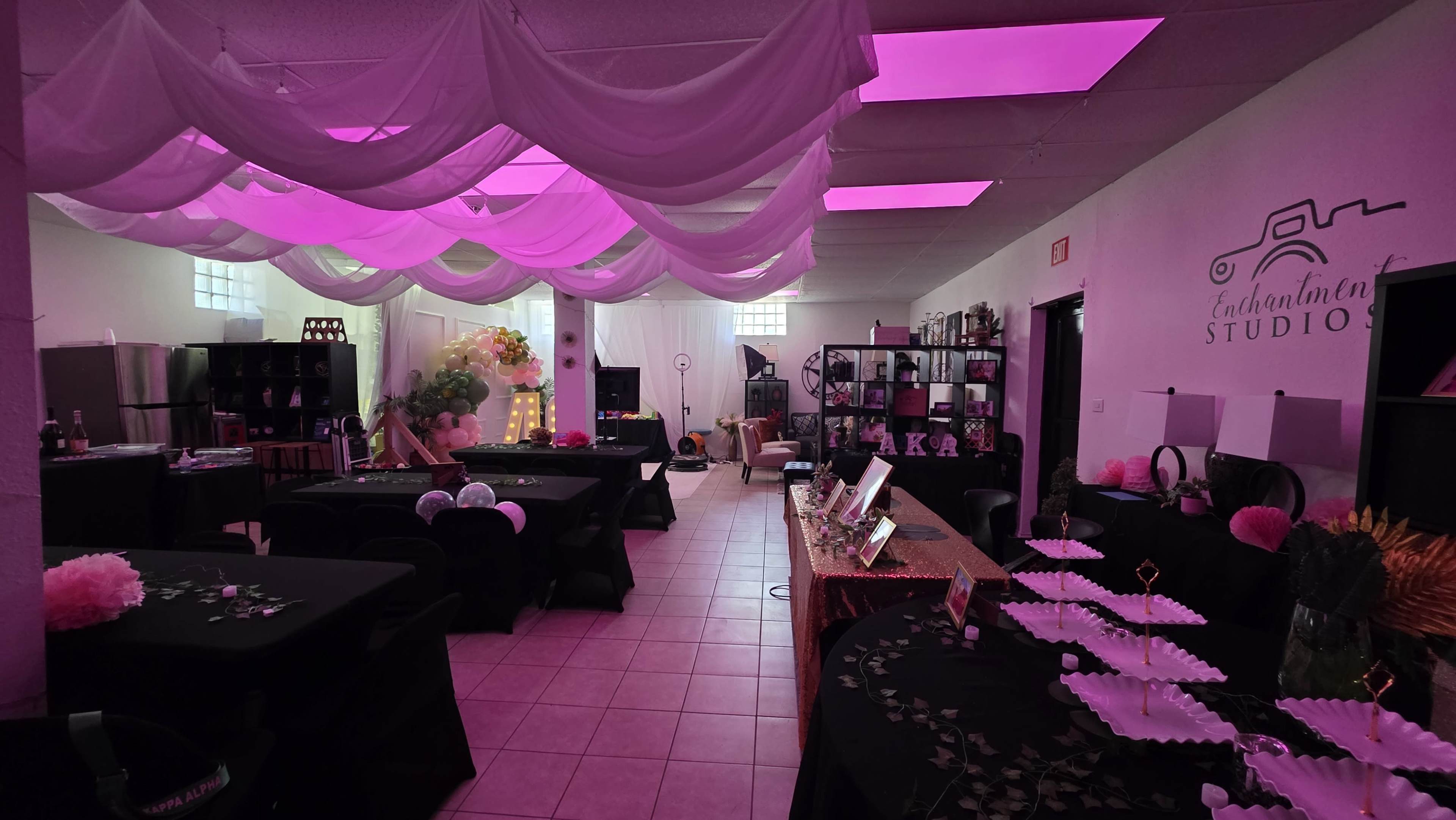 The image shows an event space adorned with draped fabric and vibrant pink lighting, featuring black tables set with decorative items and a floral arrangement at one end.