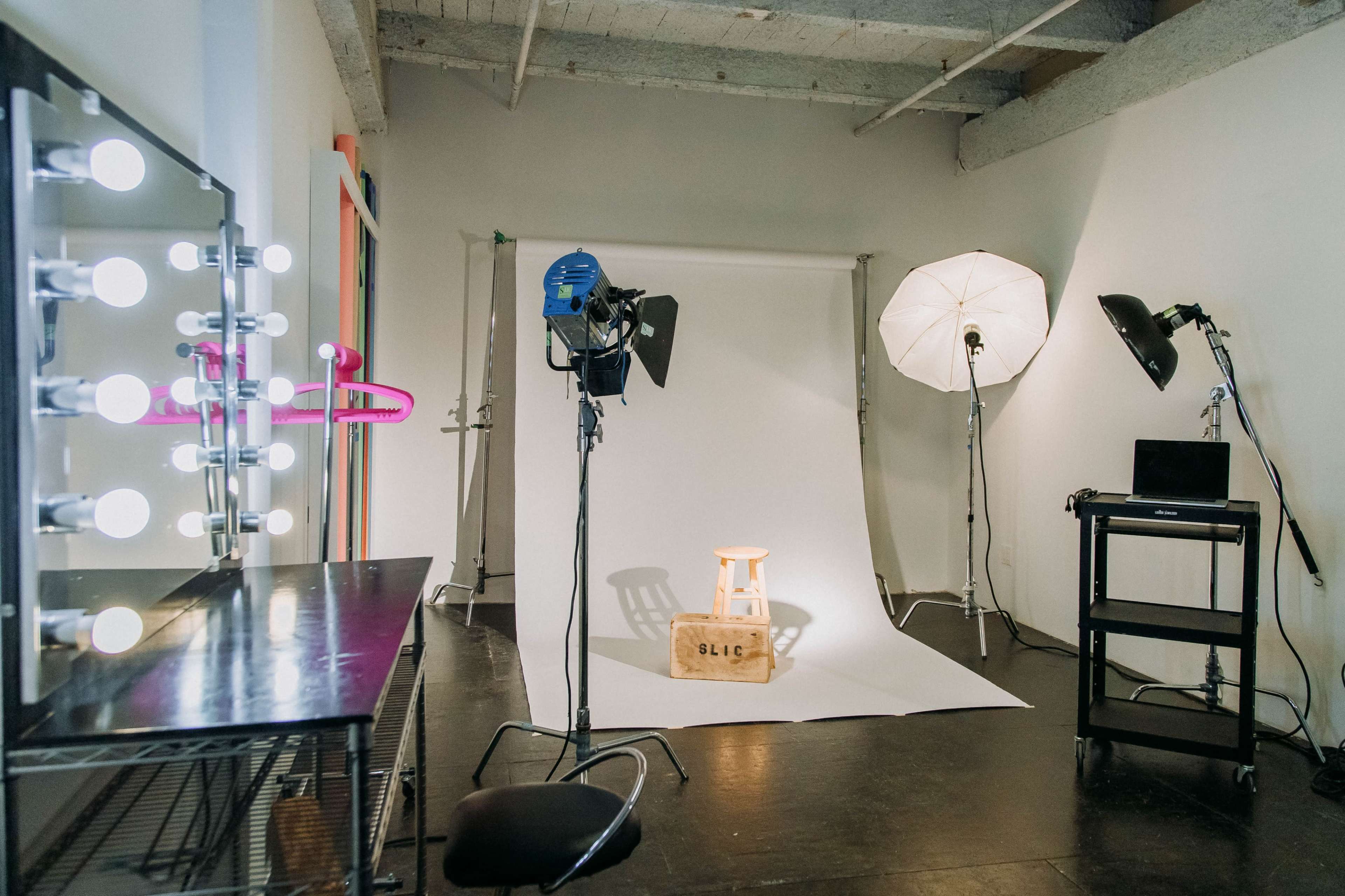 A photography studio setup with a stool on a white backdrop, lighting equipment, and a vanity mirror with bulbs.