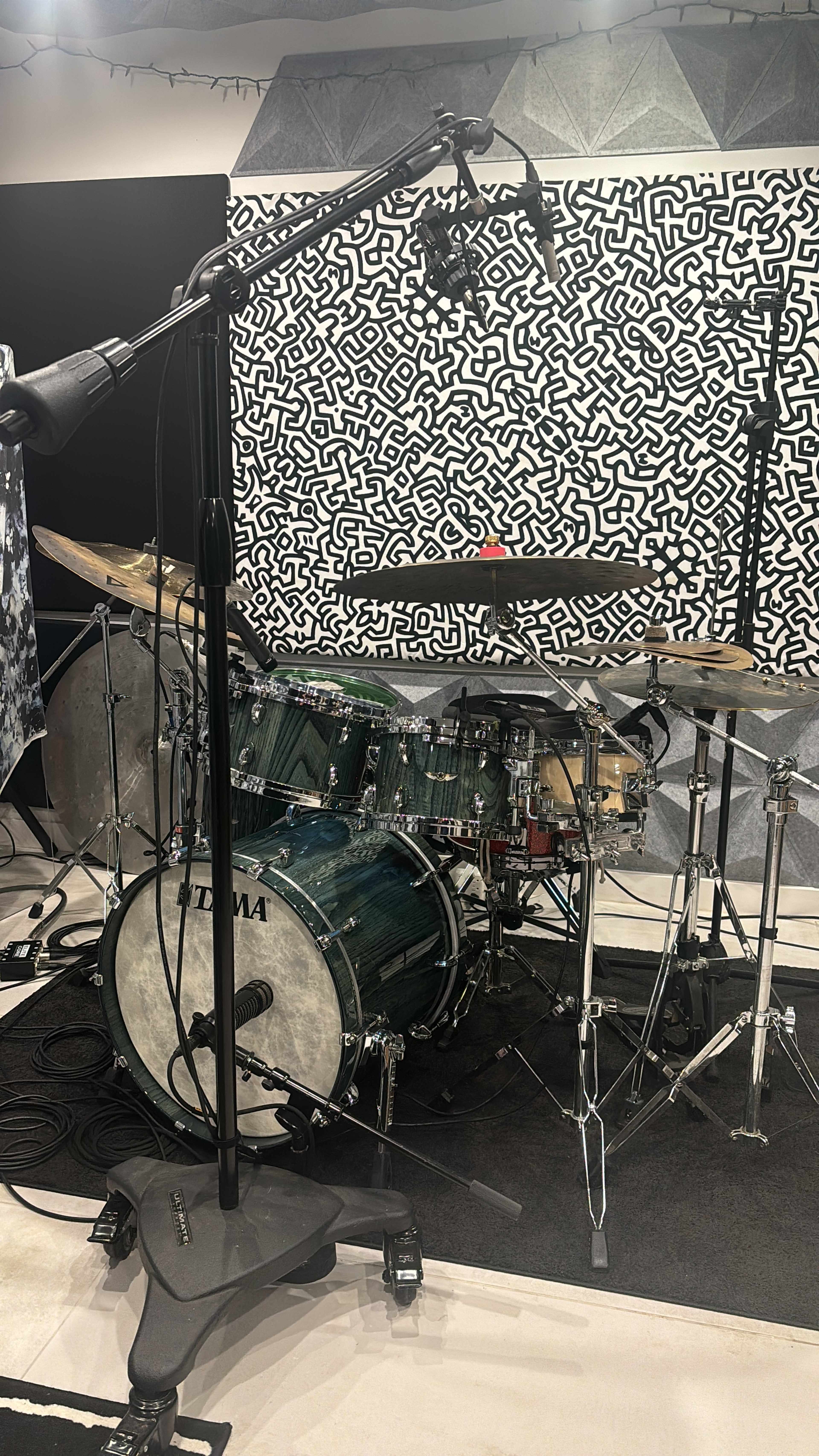 The image shows a drum set positioned in a studio with a patterned backdrop and microphone setup.