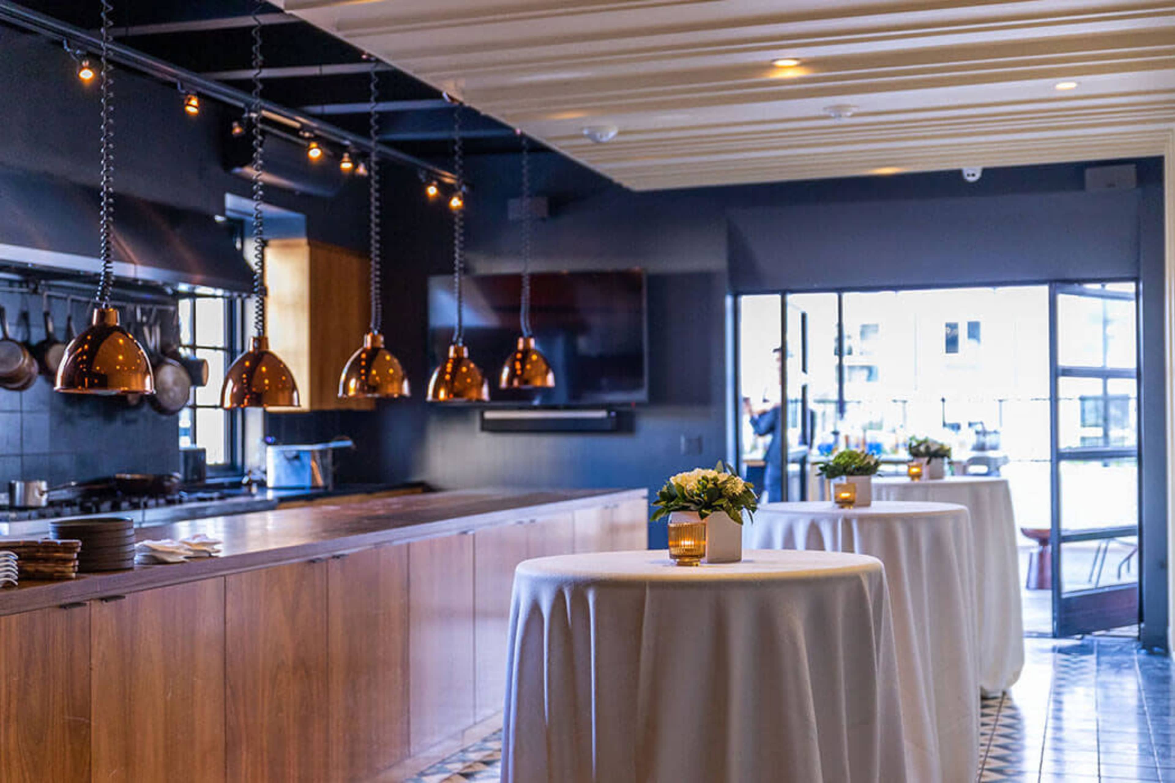 The image shows a modern restaurant interior with a long wooden counter, round tables covered in white tablecloths, and pendant lights hanging from the ceiling.