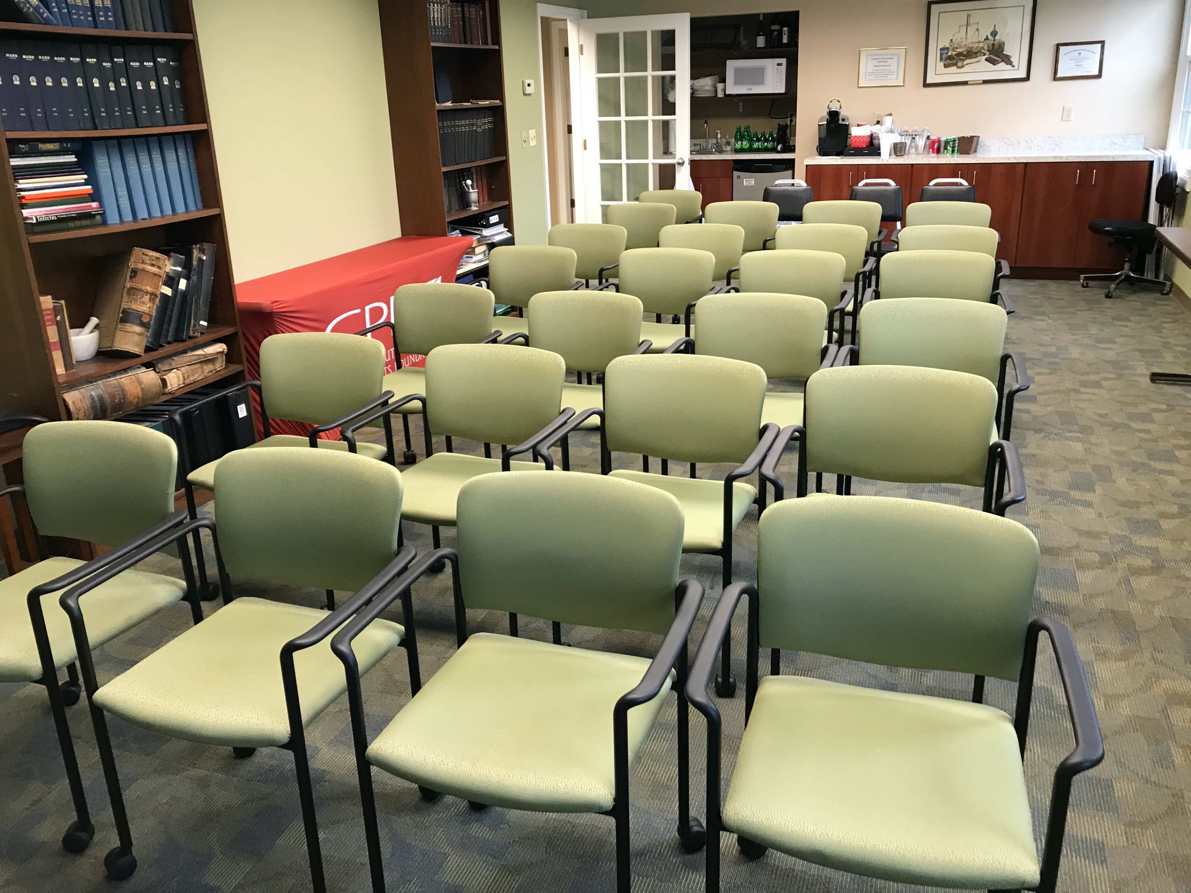 A row of green chairs is arranged in a meeting room with bookshelves and a counter in the background.