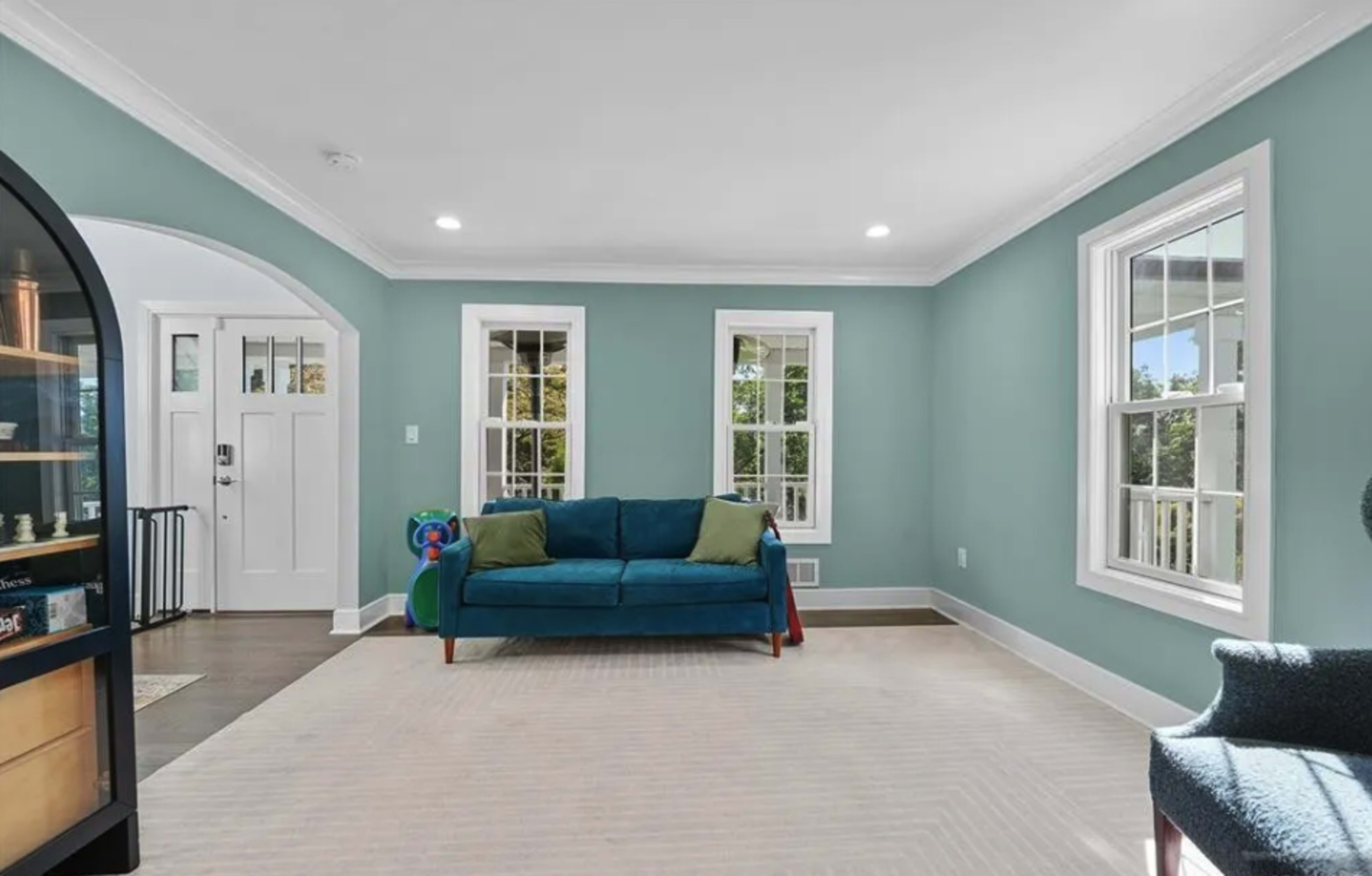 The room features a teal wall, a blue sofa with green cushions, and large windows allowing natural light to enter.