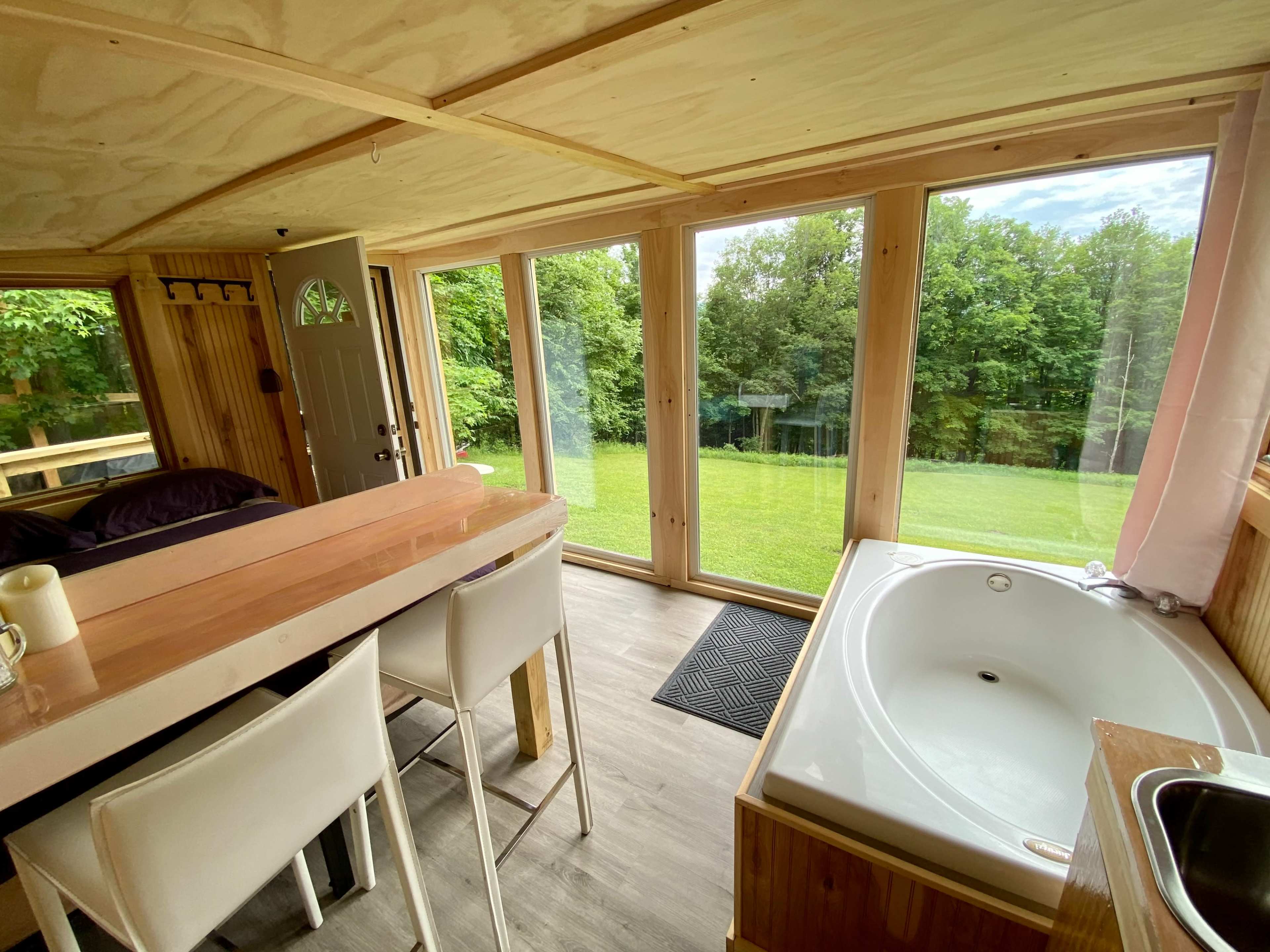 The interior of a cabin features a wood-paneled bar area with seating, a large window overlooking a grassy lawn, and a bathtub beside a sink.