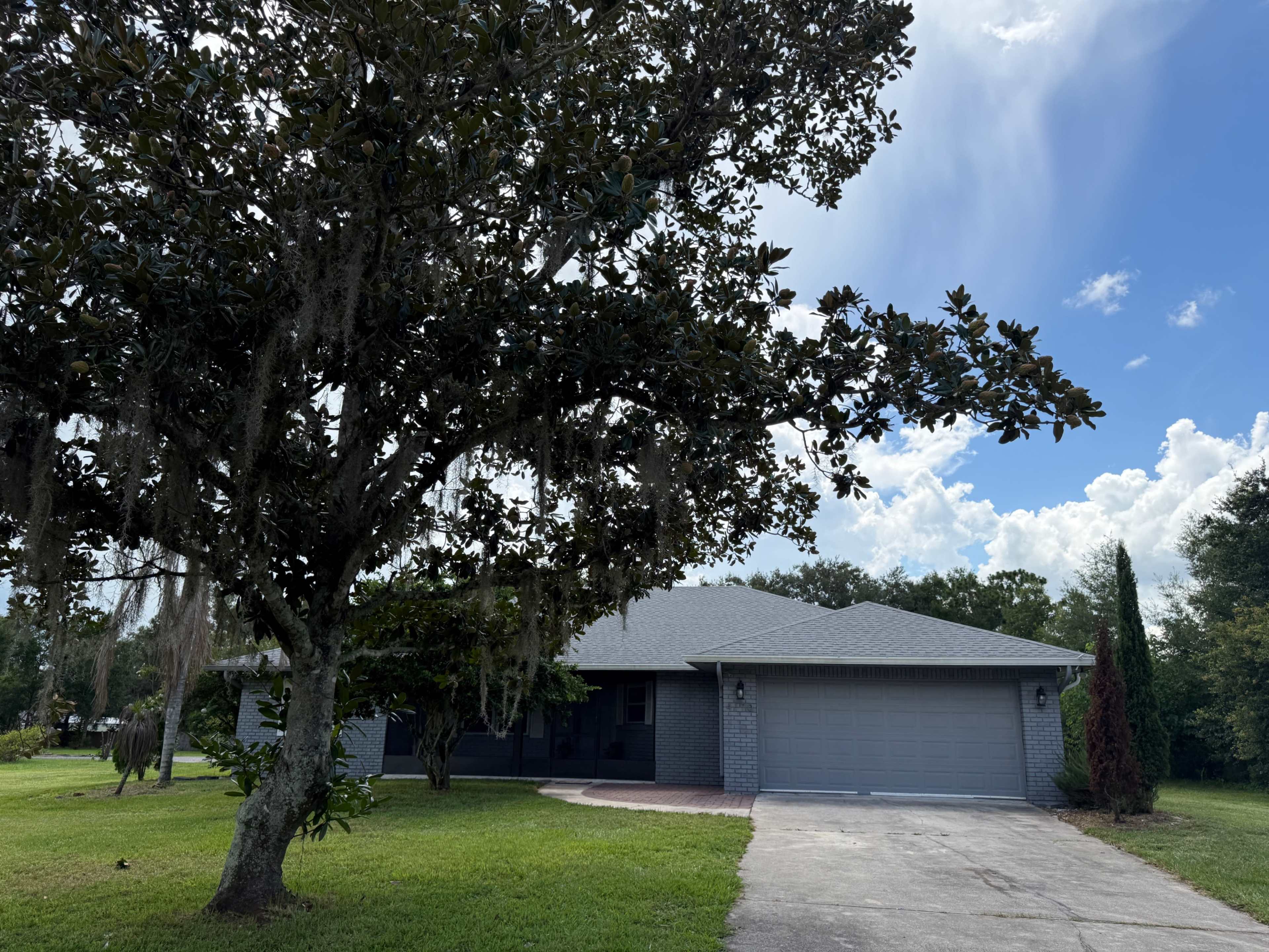 A single-story gray house with a two-car garage is situated under a large tree, surrounded by green grass and a blue sky with scattered clouds.