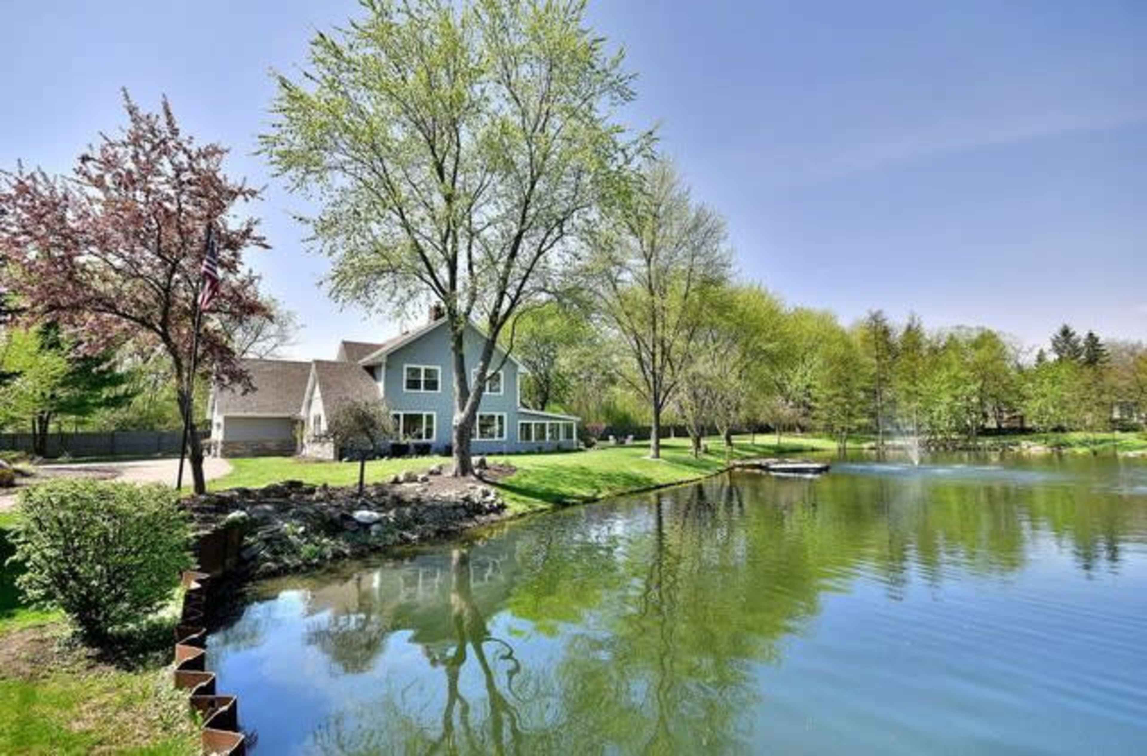 A house with large windows stands near a calm pond surrounded by green trees and landscaping.