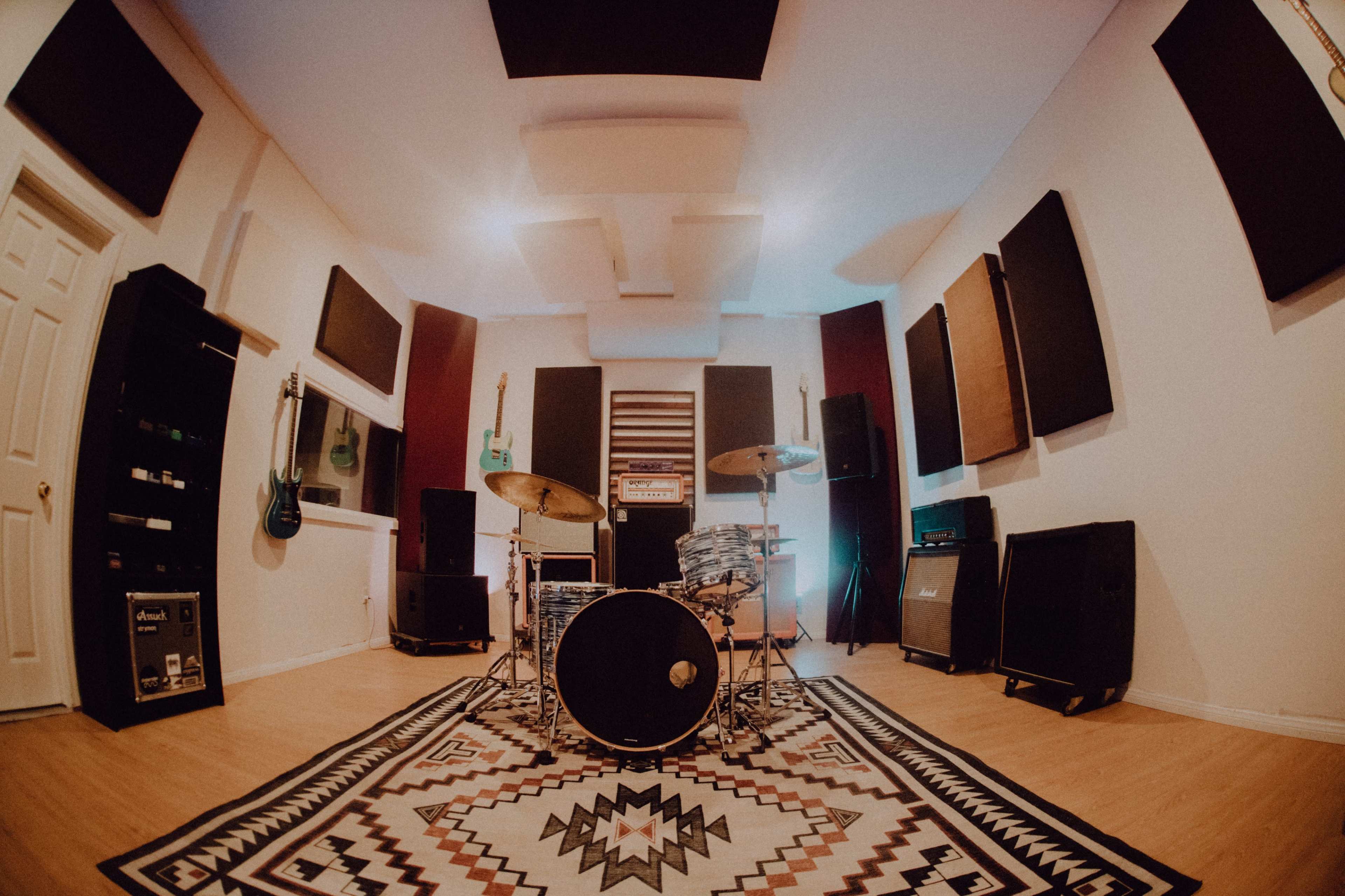 The image shows a music studio with a drum set in the center, surrounded by various guitars on the walls and soundproofing panels.