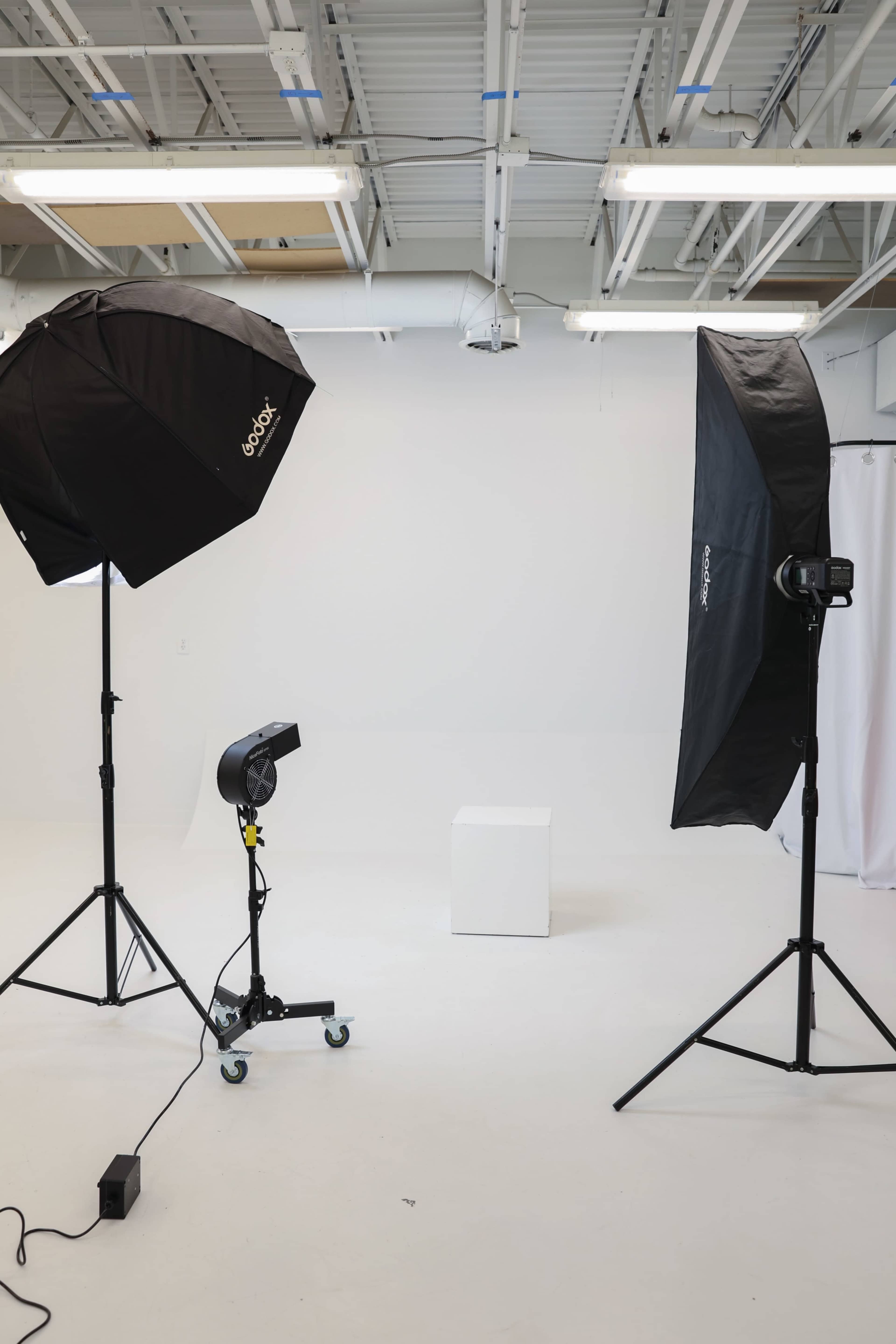 A photography studio with two softbox lights and a white pedestal on a plain, light-colored floor.