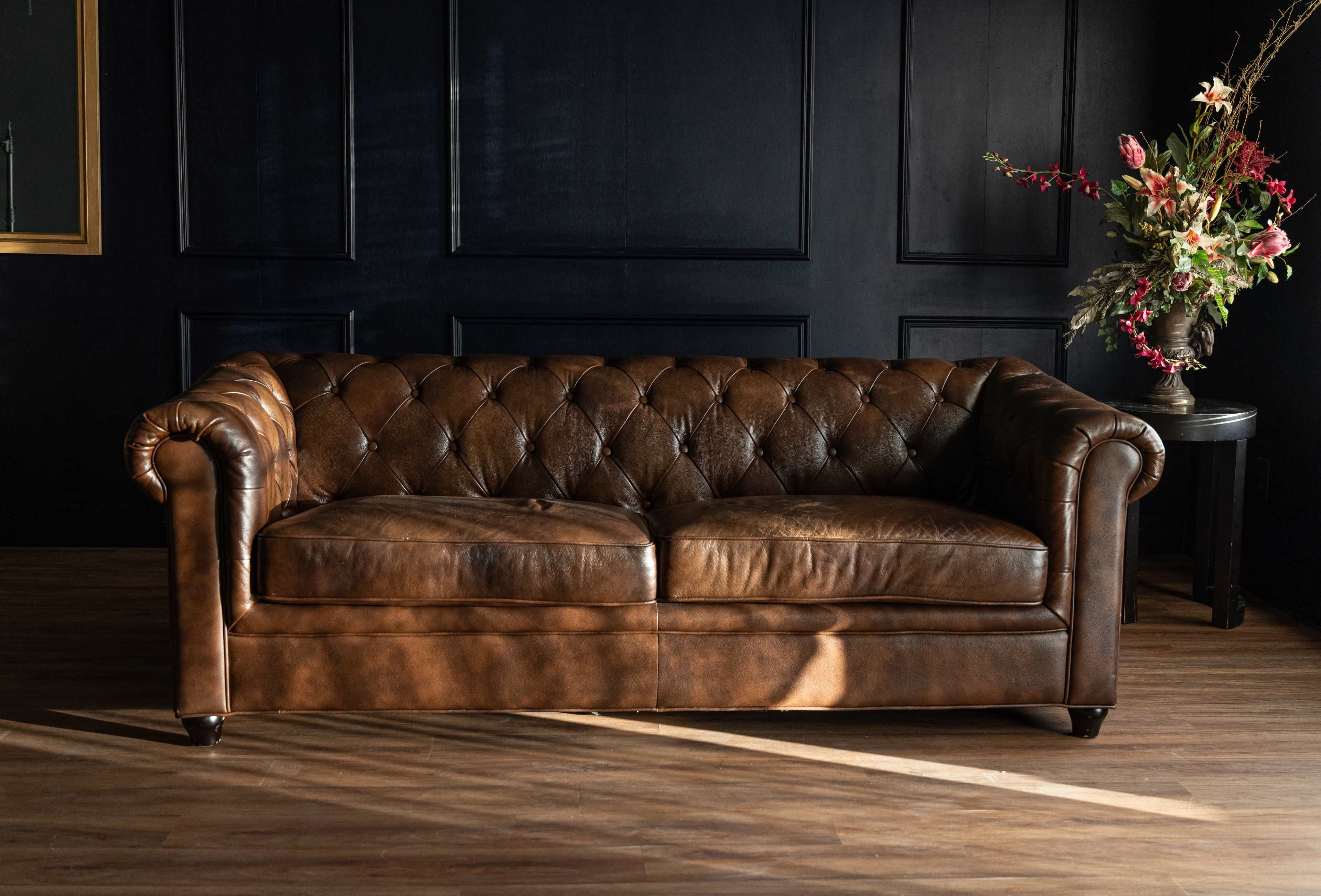 A brown leather Chesterfield sofa sits against a dark wall, illuminated by natural light, with a floral arrangement nearby.