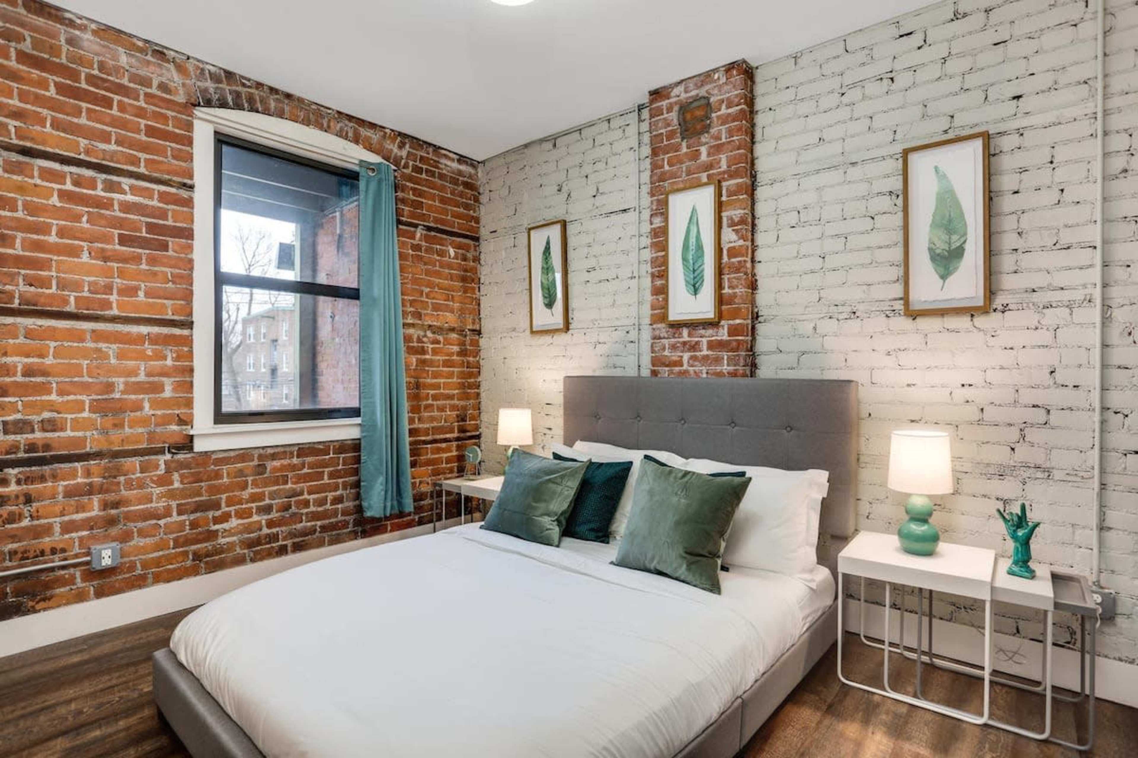 A bedroom with exposed brick walls, a gray upholstered bed, and two bedside tables with lamps.