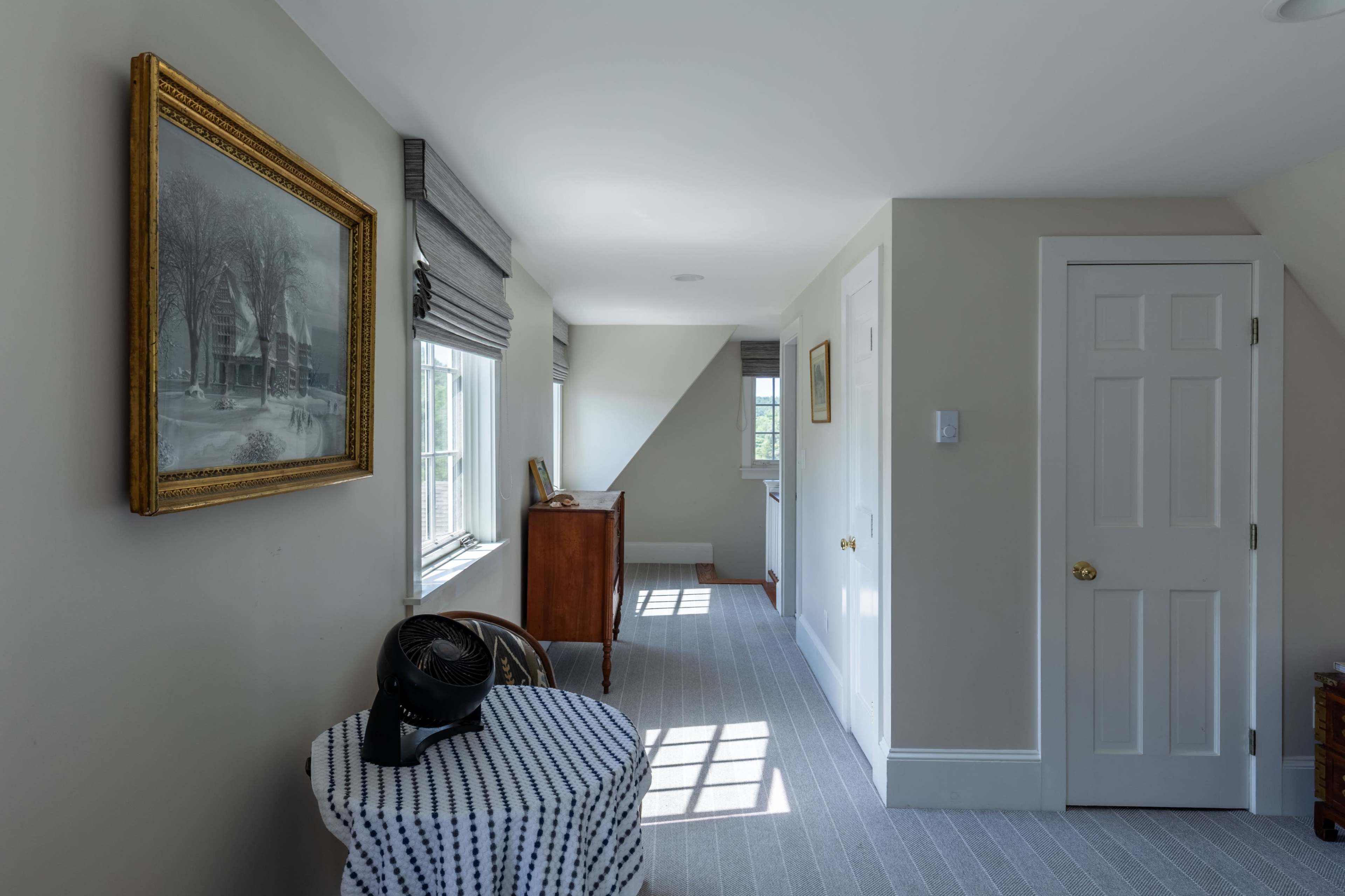 The image shows a well-lit hallway featuring a framed painting on the wall, a wooden dresser, and a closed door, with sunlight streaming through the windows.