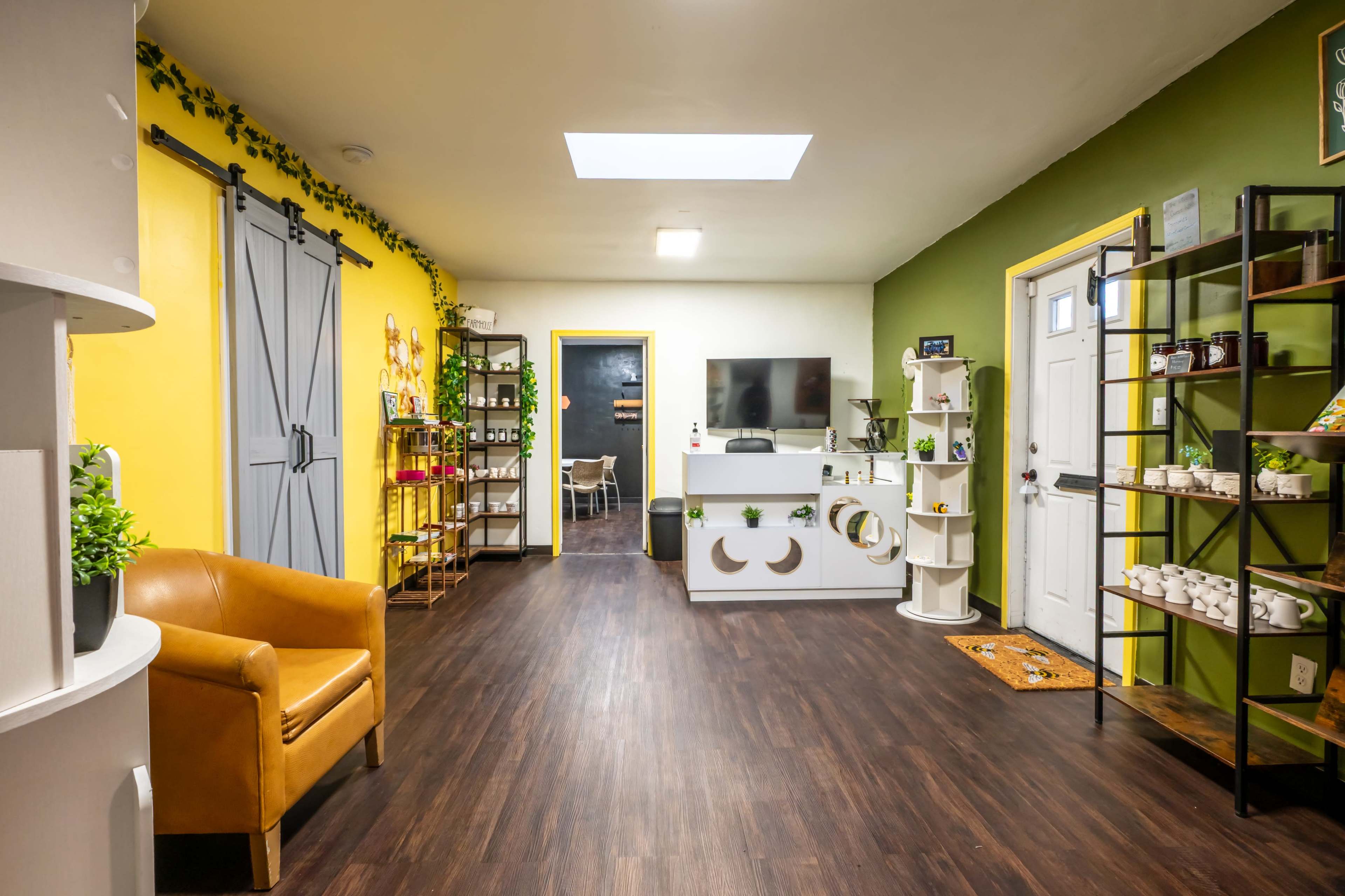 The image shows a brightly lit interior of a shop featuring green and yellow walls, a TV setup, shelves with products, and a cozy seating area with an orange armchair.
