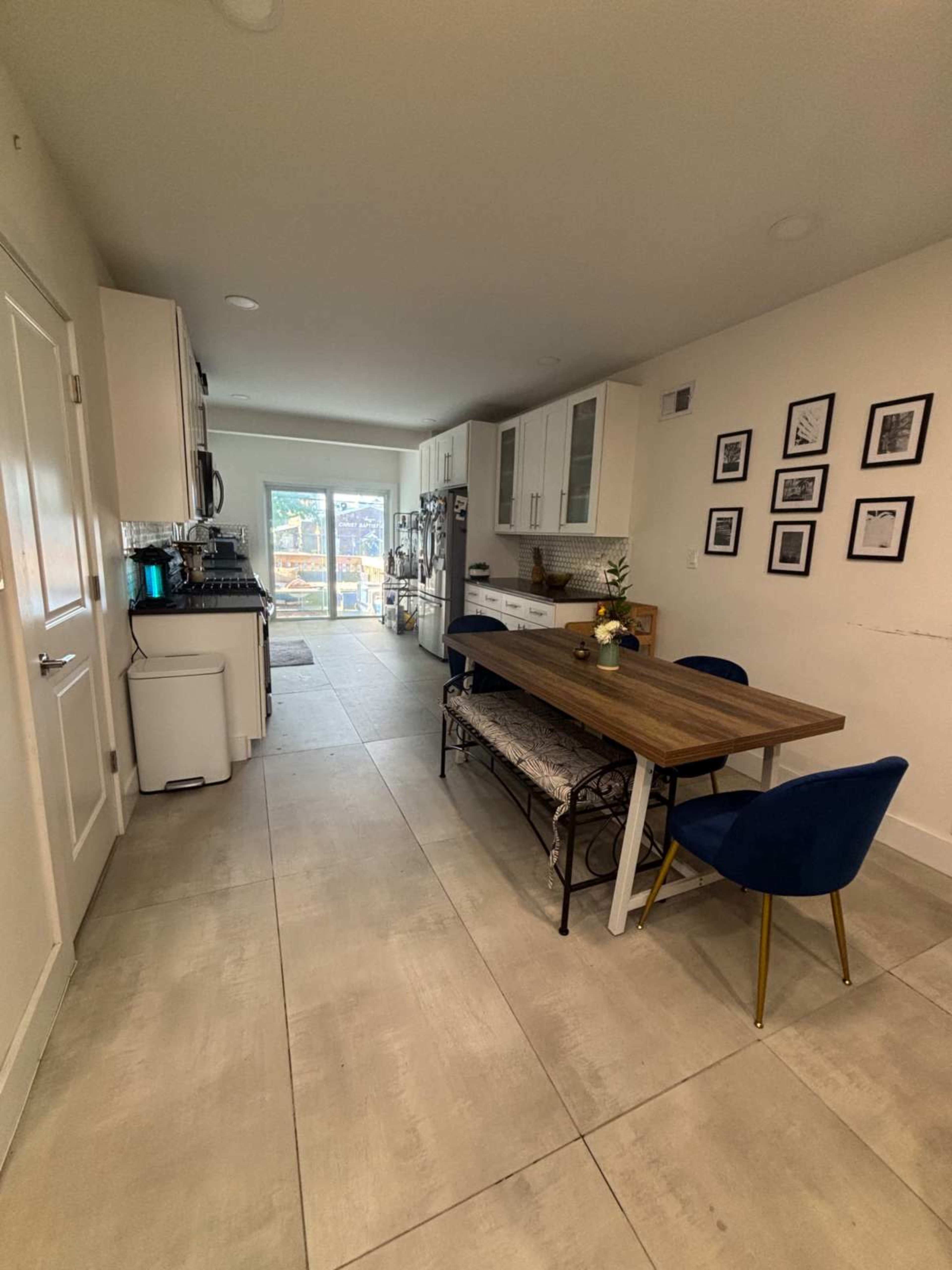 A modern kitchen features a wooden dining table with upholstered chairs, stainless steel appliances, and large windows allowing natural light.